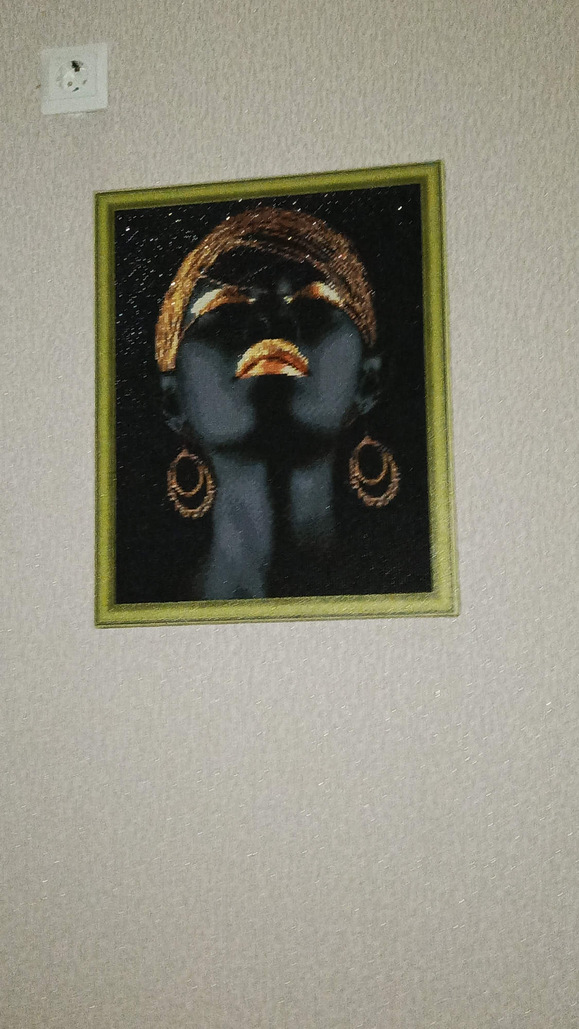 A framed artwork depicting a woman's face, painted in dark tones with gold accents, hangs on a textured, light-colored wall. The artwork is square and bordered by a thin, light green frame. The woman's eyes are closed, and her lips are parted, painted in a metallic gold. She wears gold hoop earrings and a gold headpiece. To the left of the artwork, a white electrical outlet is mounted on the wall.