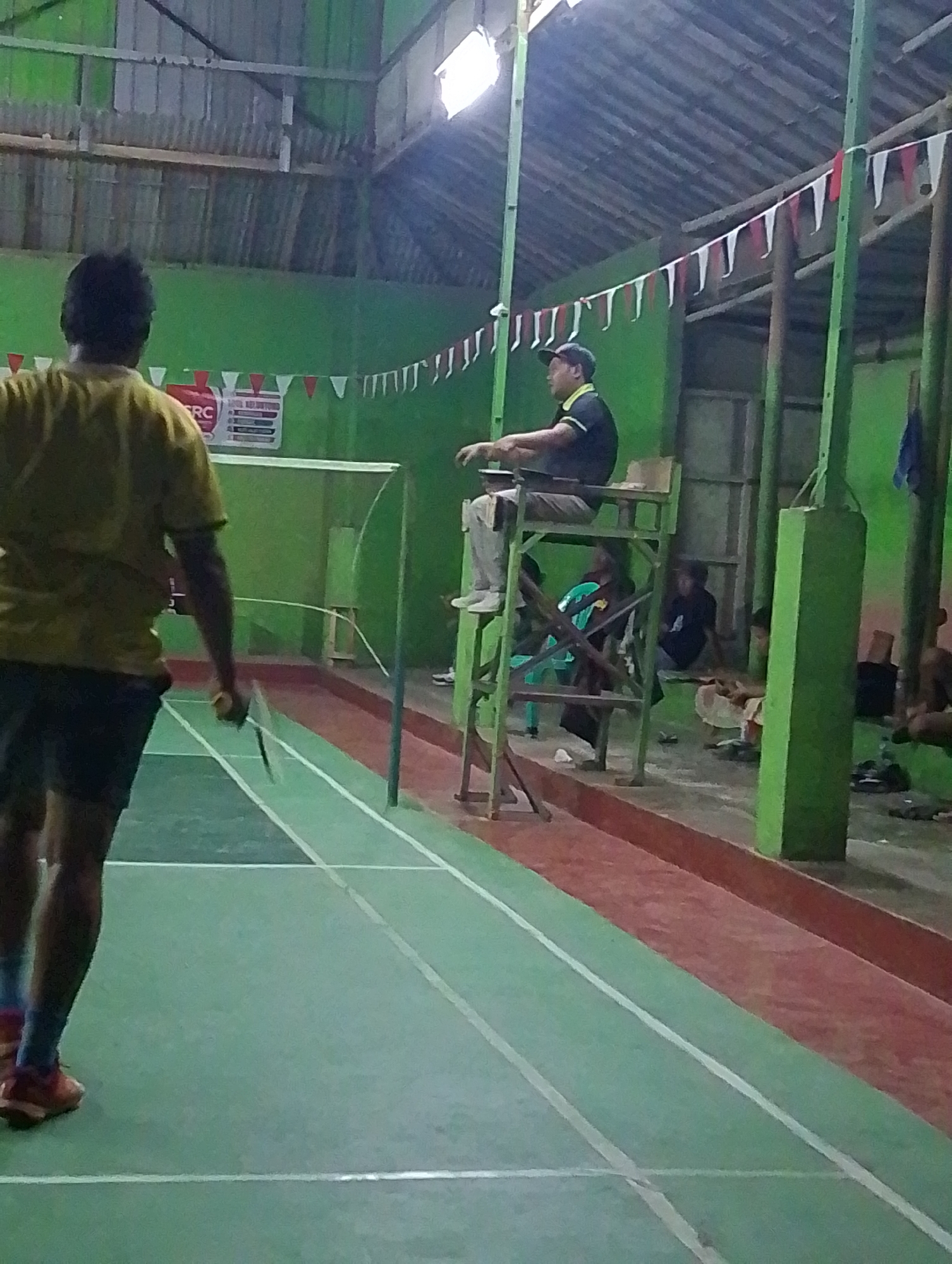 The image depicts a badminton court inside a building with green walls. A man in a yellow shirt and black shorts is on the court, holding a badminton racket. He is facing away from the camera. In the background, a person wearing a black cap and black shirt is seated on a high chair, appearing to be an umpire or referee. Several other individuals are seated or standing in the background, some obscured by shadows. The court is marked with white lines on a green surface, with a red boundary area. Triangular flags in red and white are strung along the top of the building. The lighting is dim, suggesting indoor conditions. This scene is likely taking place in Adiraja, Indonesia, a location known for its badminton courts and community sports facilities.
