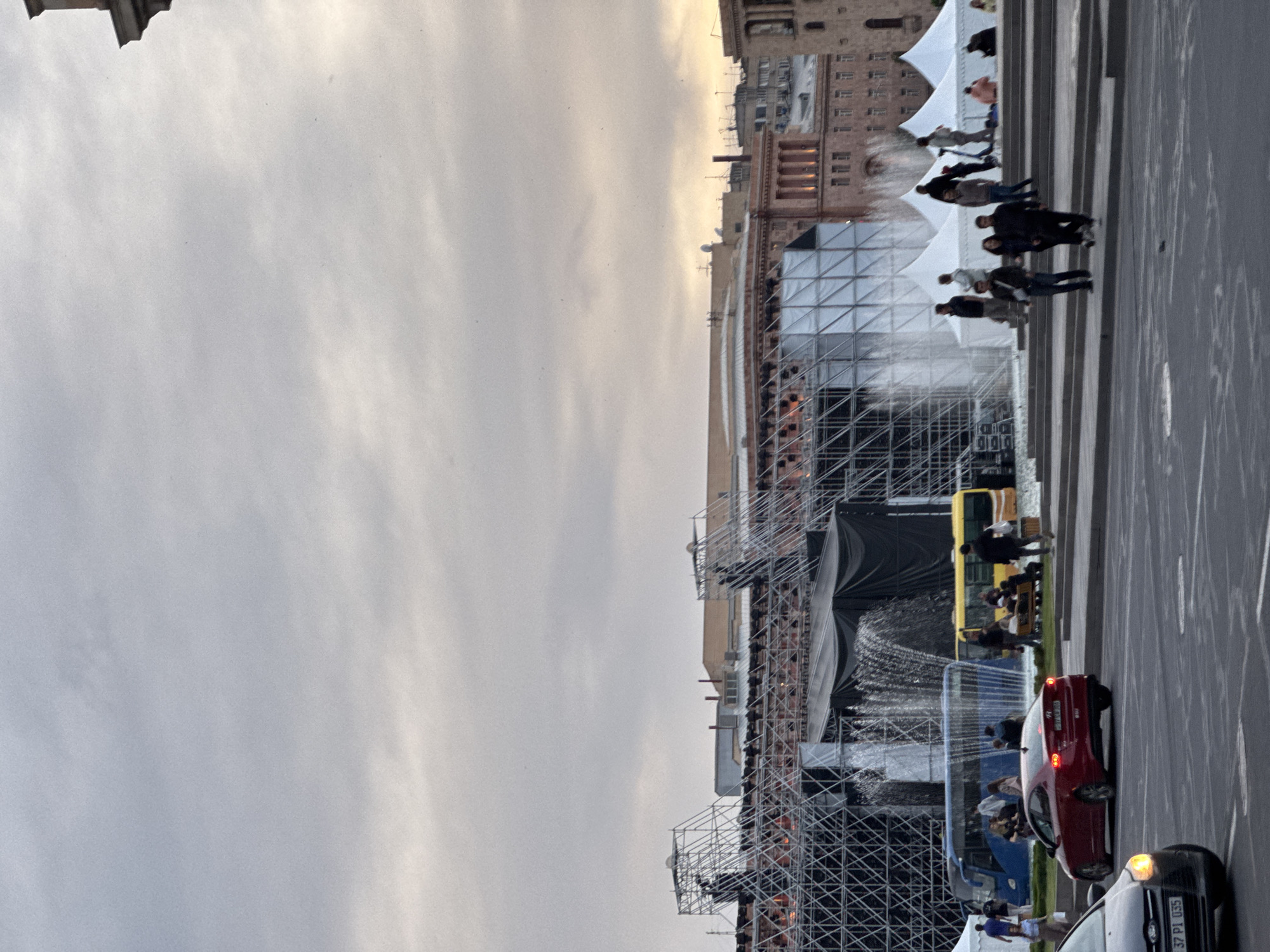 The image depicts a street scene in Yerevan, Armenia, likely in Republic Square, with a large stage structure under construction. The sky is overcast, casting a diffuse light over the scene. The architecture in the background features light-colored stone buildings with symmetrical facades. In the foreground, scaffolding supports the stage, and a fountain is visible in front. People walk along the pedestrian areas near the stage. Vehicles, including cars and a bus, are present on the street.