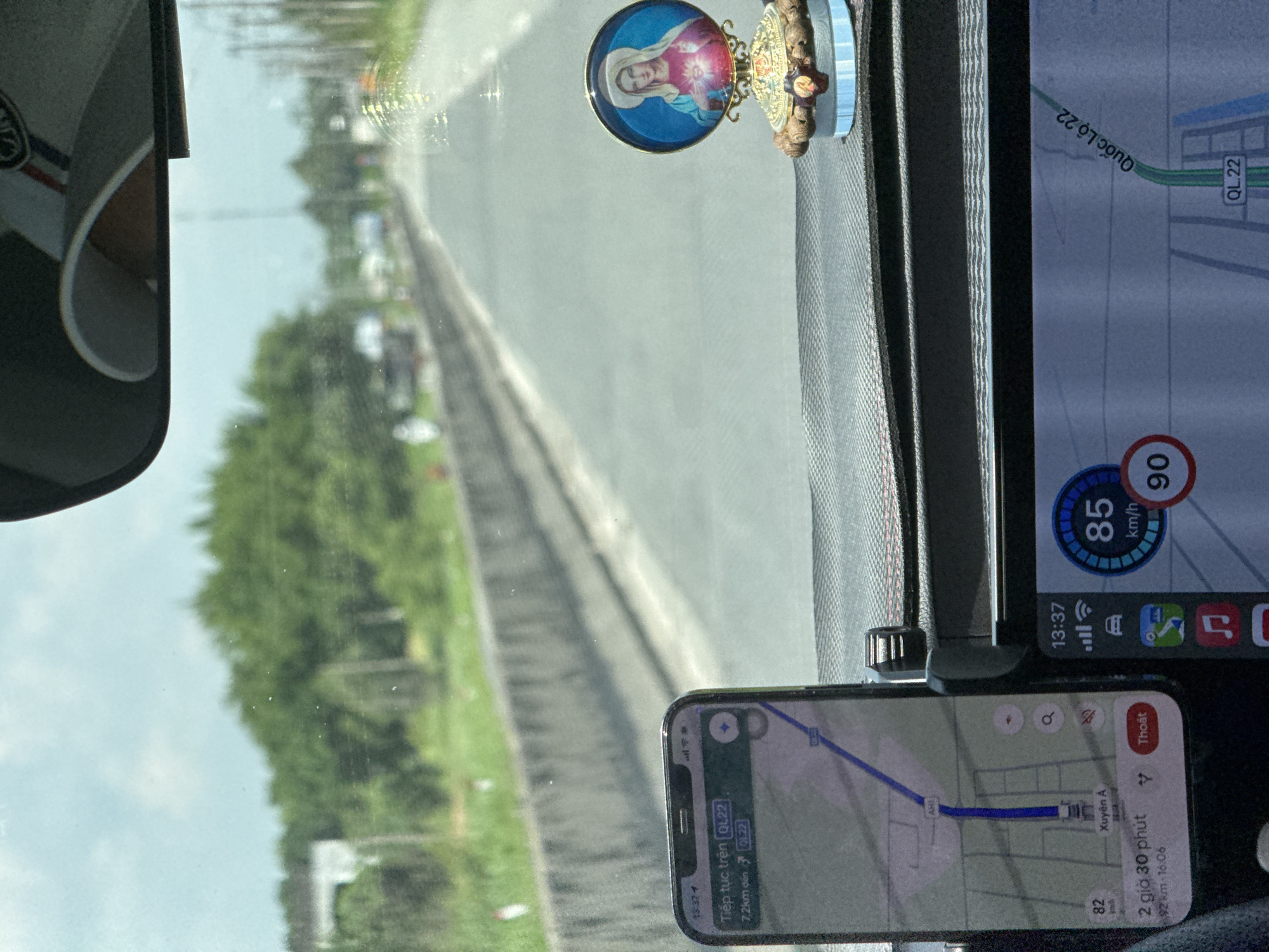 The image shows the interior of a vehicle driving on a road in Ben Cau, Vietnam. The dashboard displays a smartphone with navigation instructions and a digital speedometer showing a speed of 85 km/h. A small figurine of the Virgin Mary is visible on the dashboard. The road is lined with trees and a concrete barrier. The sky is overcast. The navigation on the phone indicates the route is on Quốc Lộ 22.