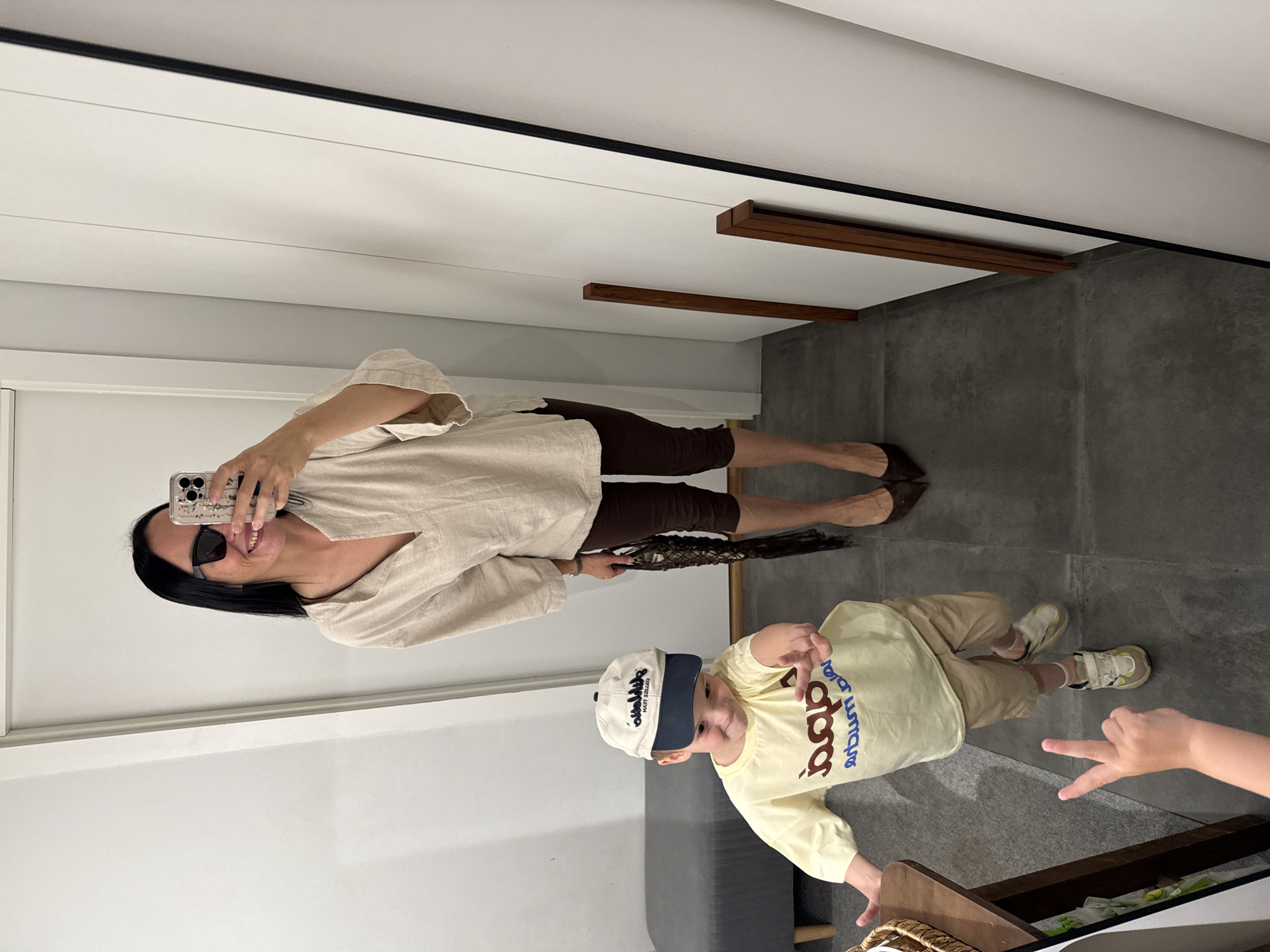 In a room with light gray walls and a dark tiled floor, a woman wearing sunglasses and a light brown shirt takes a mirror selfie. She is smiling. Beside her, a young child wearing a yellow long-sleeved shirt, tan pants, and a white and blue baseball cap looks at the camera. The child is pointing a finger and making a peace sign with their other hand. A dark brown woven object is held by the woman. The scene appears to be indoors, possibly in a changing room or a well-lit hallway. There is no indication of the time of day or weather. Text is visible on the child's shirt and hat, but it is not legible.