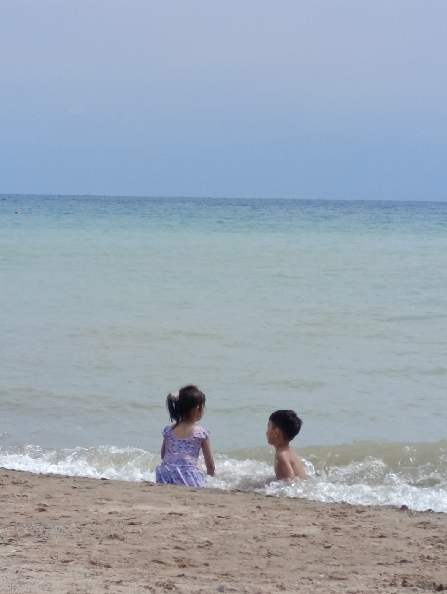 Two children, a girl and a boy, are sitting on the sandy shore of Lake Issyk-Kul in Sary-Oy, Kyrgyzstan. The girl, wearing a purple and white floral dress, is facing away from the camera, with her back to the ocean. The boy, wearing swim trunks, is also facing away from the camera, with his torso submerged in the gentle waves. The water is a light blue-green color, and the sky above is a pale blue. The sand is light brown and appears to be wet near the water's edge. The image captures a serene moment of children enjoying the beach.