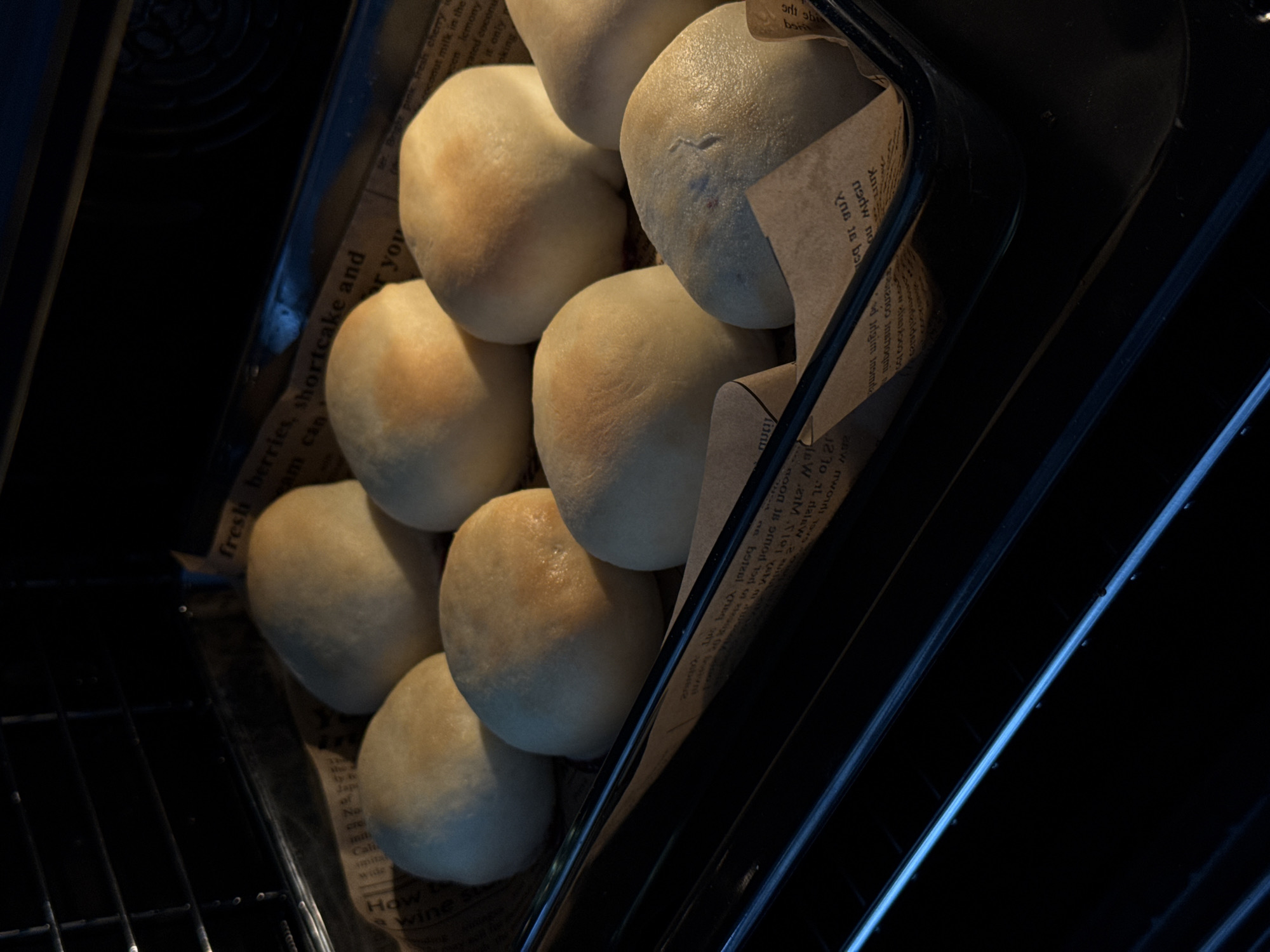 A close-up shot shows a tray of baked goods, likely buns or rolls, resting on a piece of paper with printed text that includes words like "fresh berries" and "wine," placed in what appears to be an oven. The buns have a golden-brown hue on top. The oven's interior is visible, showcasing a black wire rack and reflective elements. The setting is likely a kitchen, but the location within Moscow, Russia, is not discernible from the image.
