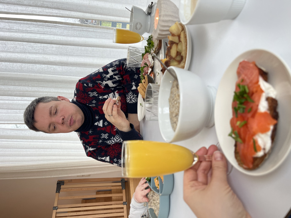 A forensic description of the scene depicts an indoor setting, likely a dining area, in the city of Gangloff, Germany. A male individual, appearing to be in his 30s or 40s, with short dark-grey hair, is seated at a white table. He wears a dark blue turtleneck sweater patterned with red and white reindeer, snowflakes, and Christmas trees. A silver ring is visible on his left ring finger. His gaze is directed downwards as he holds a small piece of food to his mouth with his right hand.
The table is laden with various breakfast or brunch items. In the foreground, a human hand holds a fluted glass containing an orange-colored liquid, likely juice or a mimosa. Slightly out of focus in the immediate foreground is a white plate holding a slice of bread topped with a white spread, several slices of smoked salmon, and chopped green chives.
Other items on the table include:
- A large white bowl containing a grey, thick substance (possibly porridge or oatmeal) in the center.
- A small white plate with a slice of apple tart or pie.
- A concrete-like, textured candle holder with a lit candle, emitting a warm glow.
- Another fluted glass with orange liquid, similar to the one held in the foreground.
- A white bowl with an embossed pattern.
- A plate with several smaller food items, including open-faced sandwiches with various toppings (e.g., ham, greens).
- A white cup, possibly containing coffee, on the far right.
- On the left, a partial view of a small child's hand is visible next to a blue, segmented plate containing a smaller portion of the grey substance and some orange-colored food items.
The background features large windows covered with sheer, light-colored curtains, providing diffused natural light. Through a gap in the curtains, faint outlines of outdoor foliage are discernible. A wooden safety gate or barrier is visible on the left side of the room. The walls are light-colored.