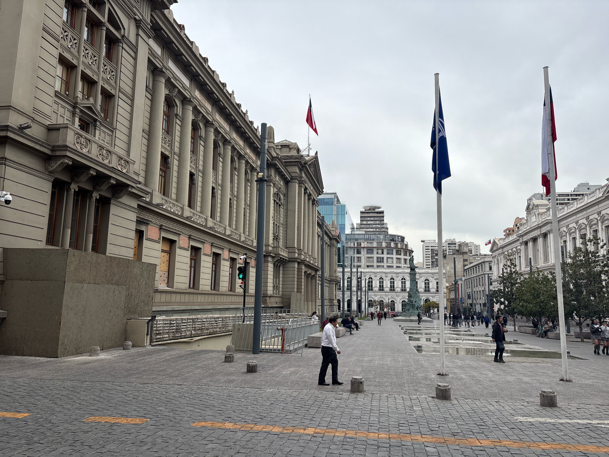 The image depicts a street scene in Santiago, Chile, likely Plaza de Armas, given the architectural style of the surrounding buildings and the presence of Chilean flags. In the foreground, a man in a white shirt and black pants walks across a cobblestone plaza. To his left, a large, modern concrete structure partially obscures the facade of an older, neoclassical building adorned with columns and decorative cornices. A CCTV camera is visible on the building's exterior.
Further into the plaza, more people are scattered, some sitting on benches, others walking. Two prominent flagpoles stand in the center of the plaza, one flying a blue flag with a white symbol and the other flying the Chilean flag. Several other buildings, varying in architectural style and height, line the sides of the plaza, suggesting an urban environment. The sky is overcast, contributing to a muted, diffused light across the scene. The ground is paved with cobblestones, and yellow tactile paving lines are visible, indicating pedestrian pathways.
