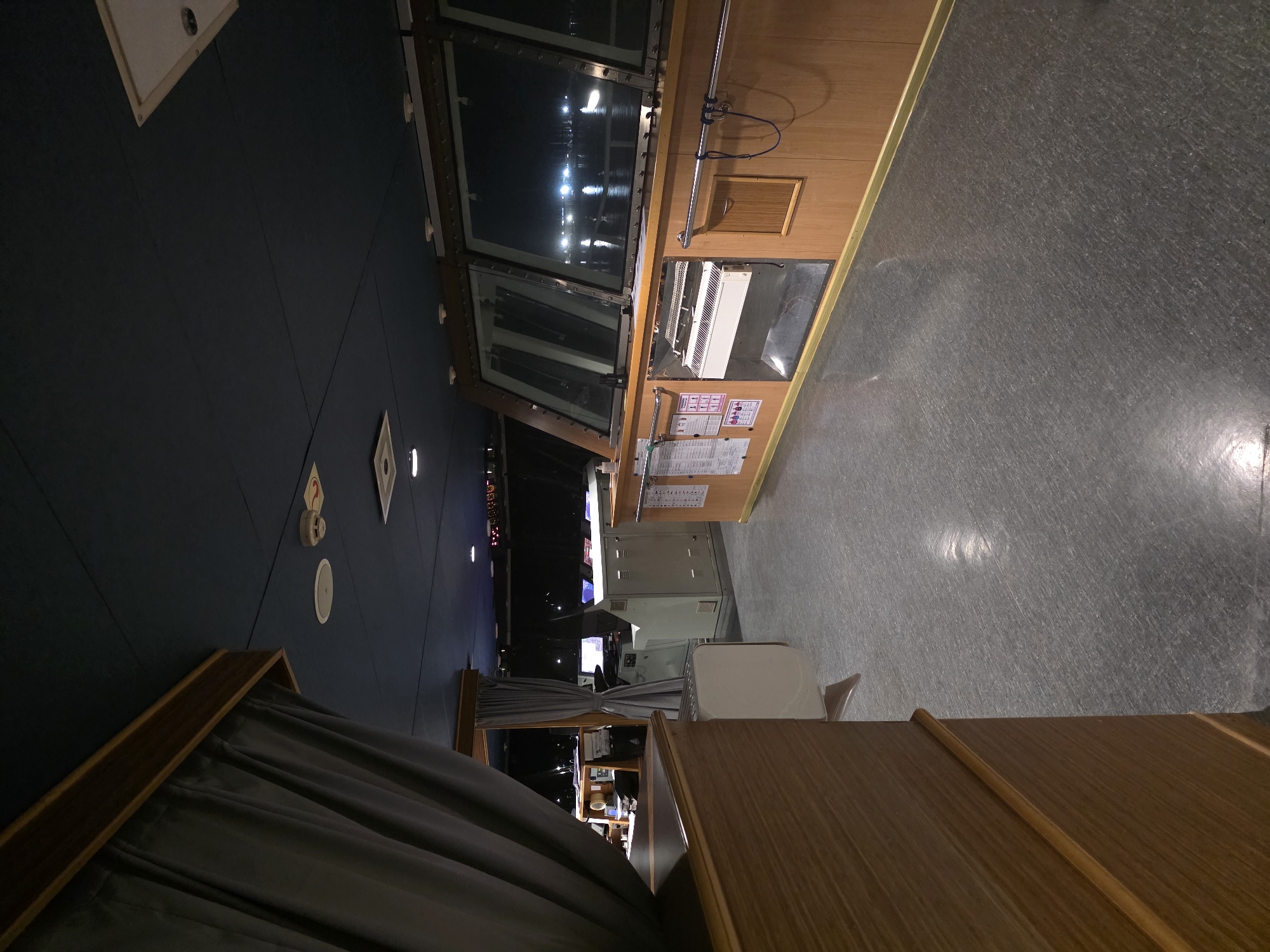 The image shows the interior of a ship's bridge at night, likely docked in Brindisi, Italy. The bridge is spacious with large windows offering a view of the dark water and distant lights of the city or harbor. The room is dimly lit, with the primary light sources appearing to be overhead ceiling lights and the reflected lights from outside.
The foreground is dominated by a wooden structure on the left, possibly a desk or console, with a grey curtain draped over it. The floor is a grey, speckled material. In the mid-ground, there is a control panel or console with various instruments and screens, some of which are illuminated. Behind this, a large white console-like structure is visible. More wooden paneling lines the walls, with metal railings and various notices or charts affixed to them. The ceiling is dark blue and appears to be made of acoustic tiles.
The overall impression is one of functional, utilitarian design, characteristic of a ship's bridge. The presence of nautical equipment and the view of the water suggest it is a working vessel. The darkness and the distant lights suggest it is late at night. The exact location within Brindisi is not discernible from the image itself, but the context points to a port or harbor setting. There are no people visible in the image.