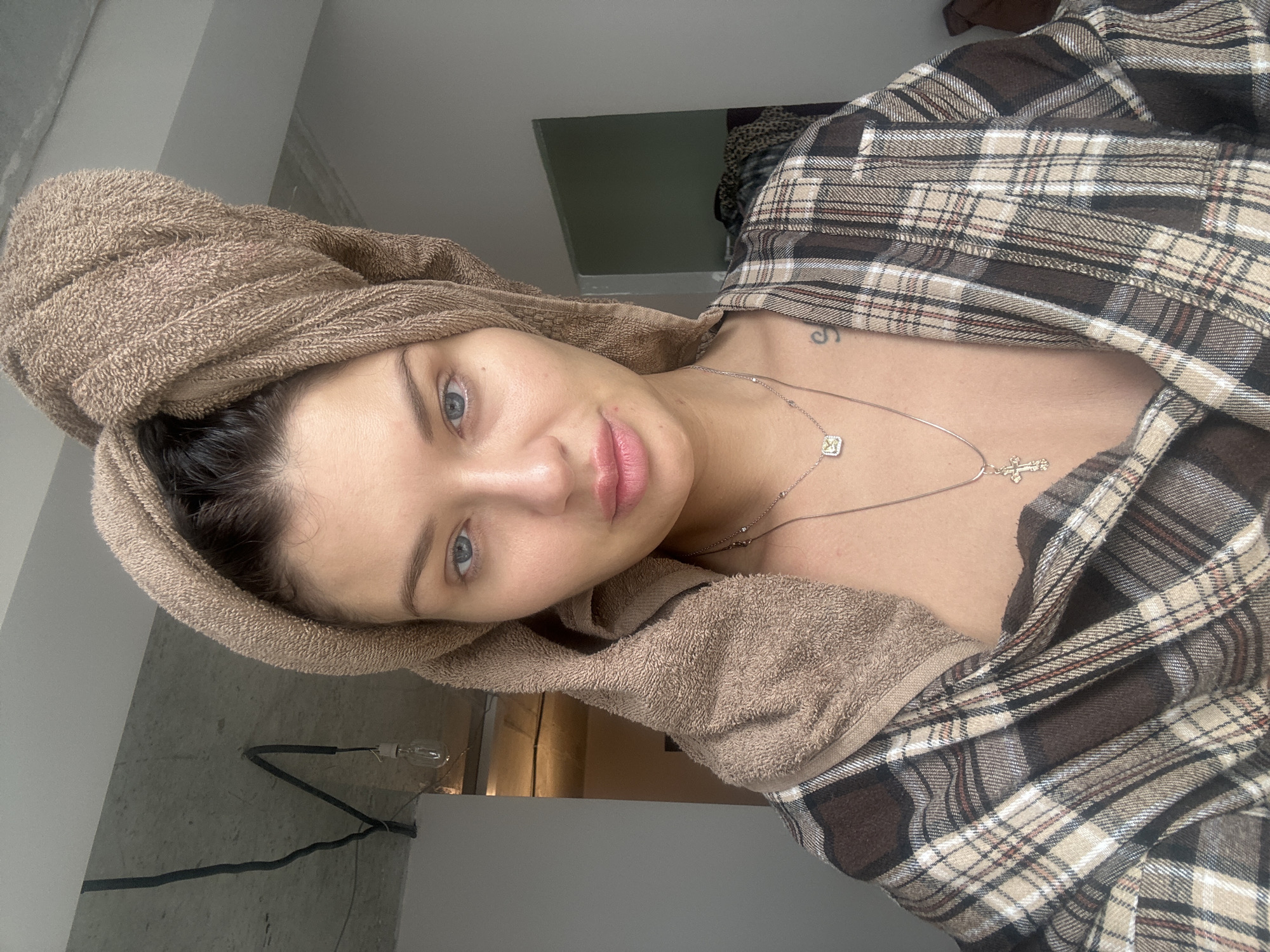 A young woman is centered in the image, wearing a brown towel wrapped around her head, and a brown and tan plaid shirt open at the neck. She has fair skin, blue eyes, and slightly parted lips. A silver necklace with two pendants, including a cross, is visible on her neck. A tattoo is slightly visible on her chest area.
The background shows a glimpse of a room interior, including a light fixture and a door. The photo appears to be taken indoors. There are no indications in the photo that would link it to Gavrilkovo, Russia.