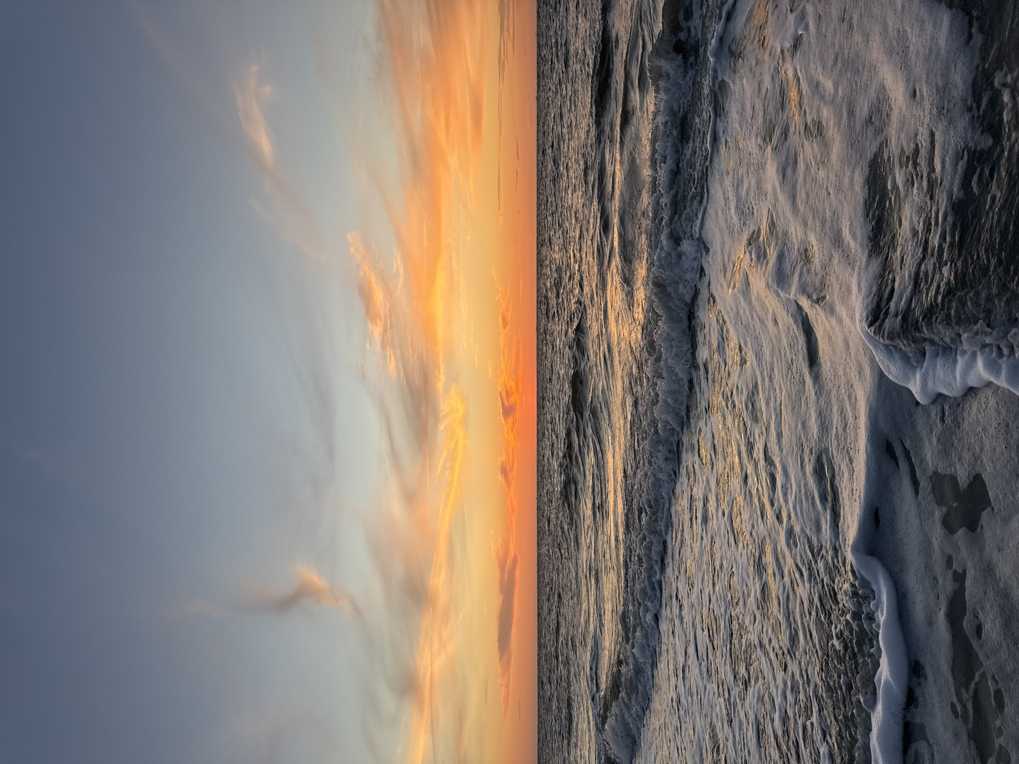 The image captures a serene sunset over the ocean on Langeoog, Germany. The sky is a gradient of soft blues at the top, transitioning into vibrant oranges and pinks as it meets the horizon, painted with wispy clouds catching the last rays of sunlight. The ocean below is calm, with gentle waves rolling towards the shore. The foreground shows the foamy edge of a wave washing onto the dark, wet sand, creating intricate patterns of white foam against the darker water. There are no people or man-made objects visible in the scene, emphasizing the natural beauty of the coastal environment. The overall impression is one of tranquility and the awe-inspiring spectacle of a sunset over the sea.