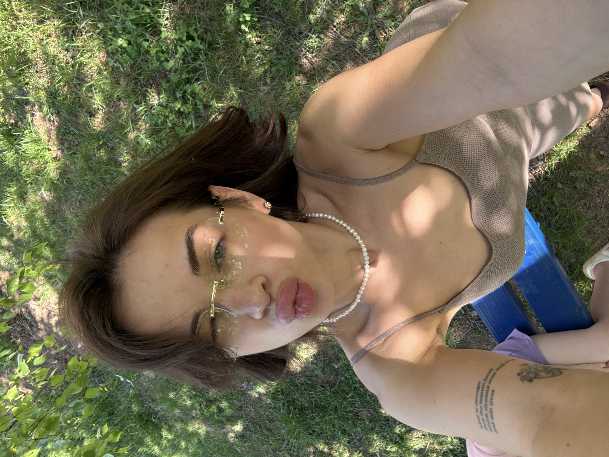 A woman takes a selfie outdoors. She is wearing a beige knit top, pearl necklace, and transparent glasses with gold frames. She is sitting on a blue bench. A child in a purple shirt is partially visible to the right of the woman. The background consists of grass and greenery. A tattoo is visible on her left arm. There is no evidence this photograph was taken in Sarapul, Russia.