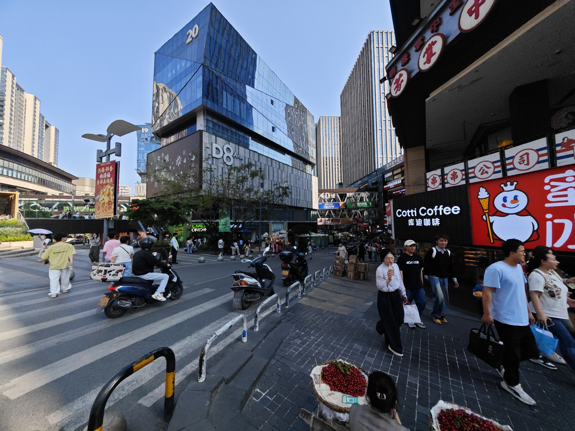 The image presents a bustling street scene in a commercial district within Yuzhong District, China, during a clear and sunny day. The setting is dominated by modern, glass-clad skyscrapers, with one large building prominently marked "20" and "D8 Ave.", and another displaying "19".
The street is active with numerous pedestrians walking on sidewalks and crossing the road via a zebra crossing. Several people are riding motorized scooters; one is captured mid-crosswalk. On the sidewalk, a vendor is selling fruit, likely cherries, from large baskets. People are seen carrying shopping bags and drinks, contributing to the busy urban atmosphere.
Visible commercial text includes "Cotti Coffee" and its Chinese equivalent "库迪咖啡", along with the distinctive snowman logo of Mixue Bingcheng. Other signs feature "热辣鲜香", "向得福", and "香辣美蛙鱼" (Spicy and delicious bullfrog fish), suggesting food establishments. Further text spotted includes "LINLI", "《手打>柠", "时代汇南", "四方朋友", "KTV", "404108", and various Chinese characters such as "掌", "芳", "公司" (Company), and "串" (Skewer) on diverse storefronts and signs. The scene vividly portrays a vibrant and active urban environment characteristic of modern Chinese cities.