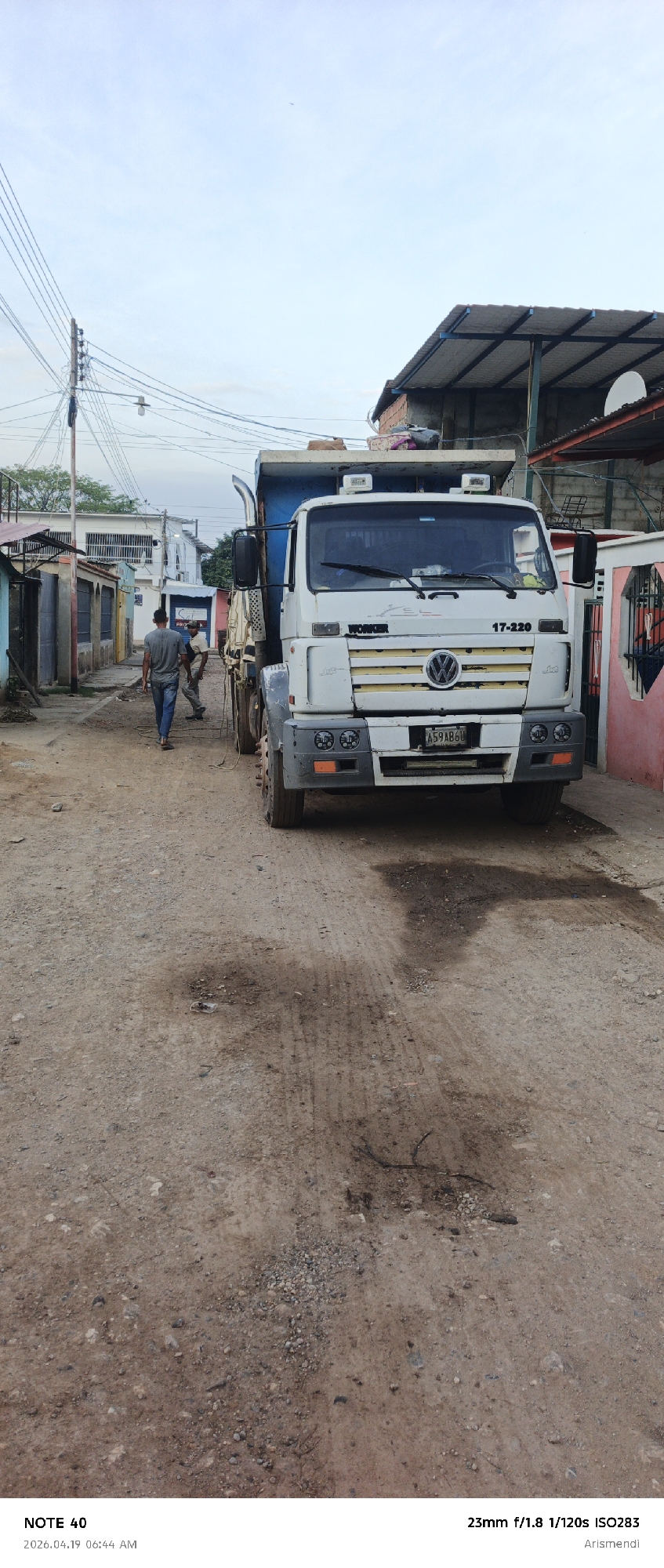 The image depicts a Volkswagen 17-220 dump truck parked on a dirt road in a residential area of Arismendi, Venezuela. Two men are walking away from the truck down the road, which is lined with buildings on either side. The sky is overcast. The truck's license plate reads "A59AB6U." The road surface is uneven and has areas with oil stains. Power lines are visible overhead. The camera settings used were 23mm f/1.8, 1/120s, and ISO283. The date and time of the photograph are 2026.04.19 06:44 AM.