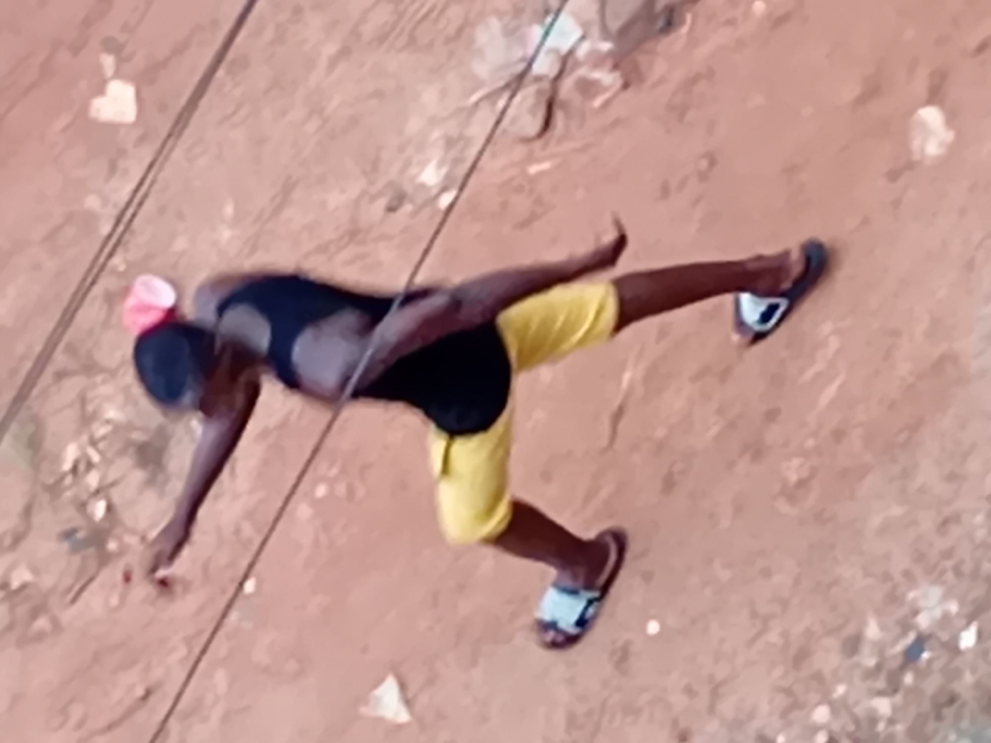 A person is captured in mid-stride on a reddish-brown dirt path in Nkpor, Nigeria. The individual, wearing a black tank top and yellow shorts, has their left leg extended forward and their right leg bent at the knee, suggesting forward motion. They are wearing blue and white flip-flops on both feet. A pink object, possibly a cap, is visible on their head. The background consists of the uneven dirt surface with scattered debris, including white paper-like items and some darker specks. Overhead, electrical wires are visible. The lighting suggests it is daytime.