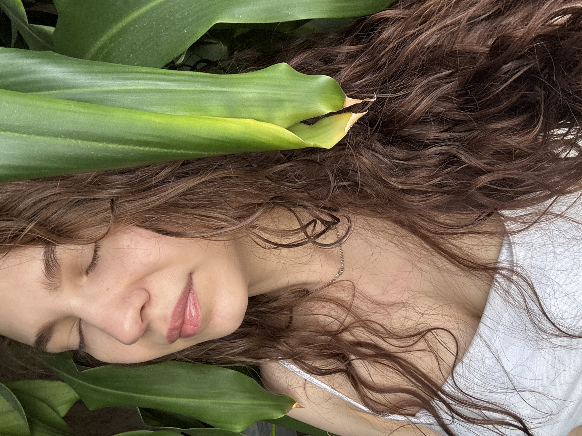 A person with closed eyes and long, brown, wavy hair is lying among large, green leaves. The person is wearing a white top and a delicate silver necklace. The image appears to be taken from above, capturing the person's face, hair, shoulder, and the surrounding greenery. The background is blurred. There is no indication of the location being Nizhnyaya Gar', Russia.