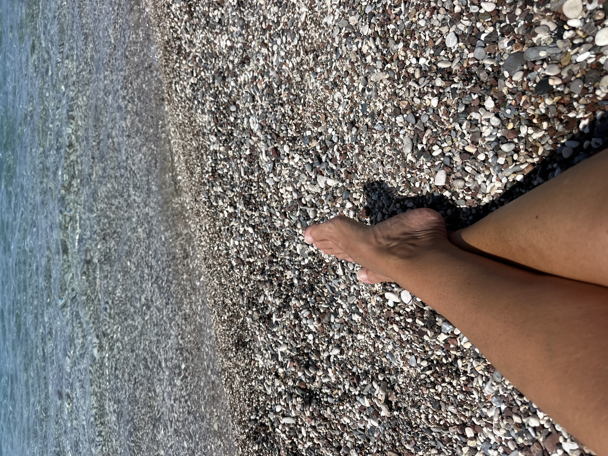 The image depicts a view from above, capturing a portion of a beach with legs extending into the frame. The right side of the image features two legs resting on a beach covered with small, smooth, multicolored pebbles. To the left, the sea is visible, its surface clear with subtle ripples. Based on the location of the image, the beach is located in Budva, Montenegro. No other individuals or objects are visible.
