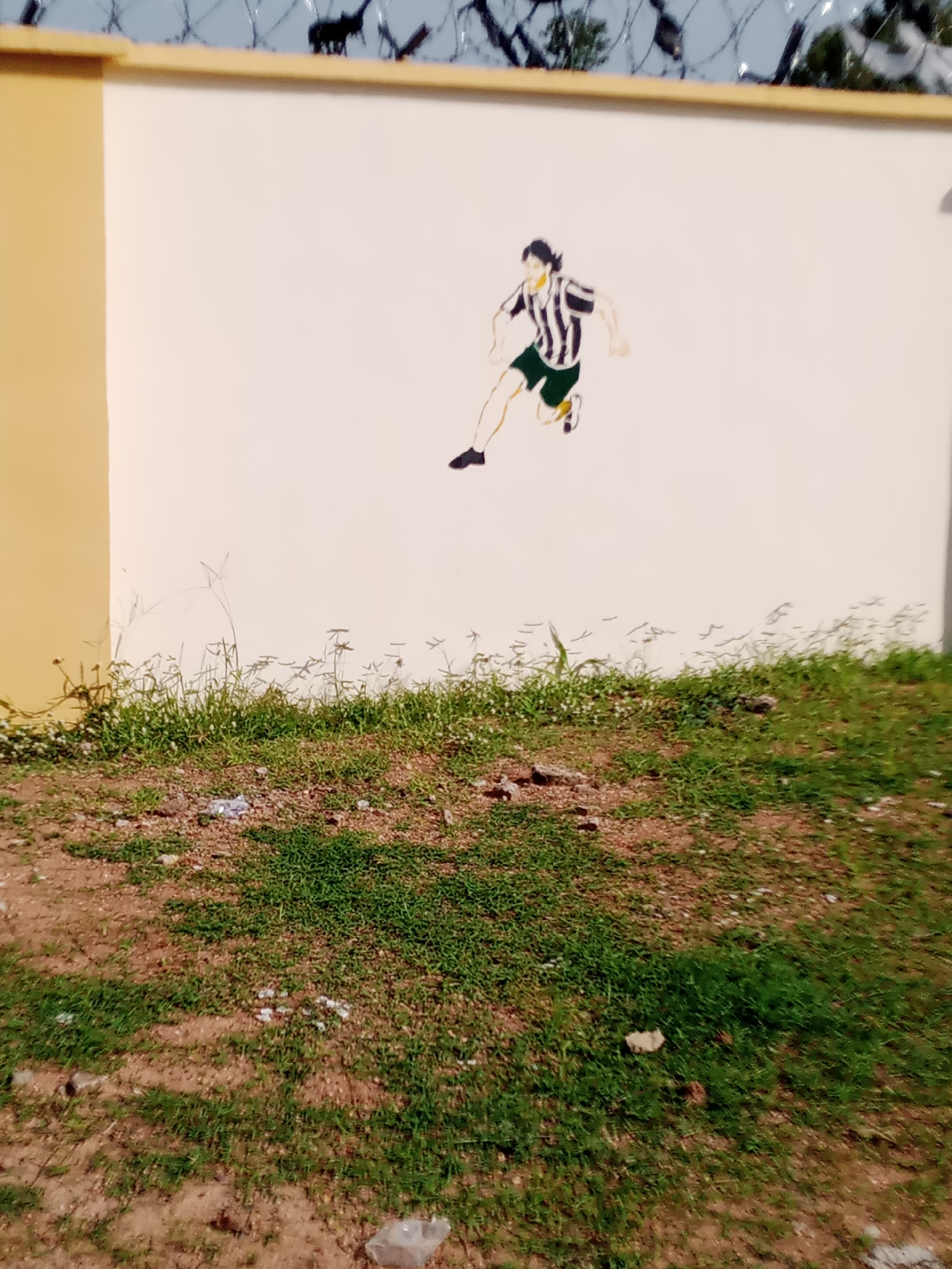 The image depicts a mural of a soccer player on a lightly colored wall, likely in Jalingo, Nigeria. The player is rendered in a stylized, cartoonish manner, wearing a striped jersey and shorts, with arms outstretched as if in motion. Above the wall, barbed wire and a fence are visible, suggesting a secure area or perimeter. The ground in front of the wall is covered with sparse grass and some litter. The overall scene appears to be an outdoor urban environment.