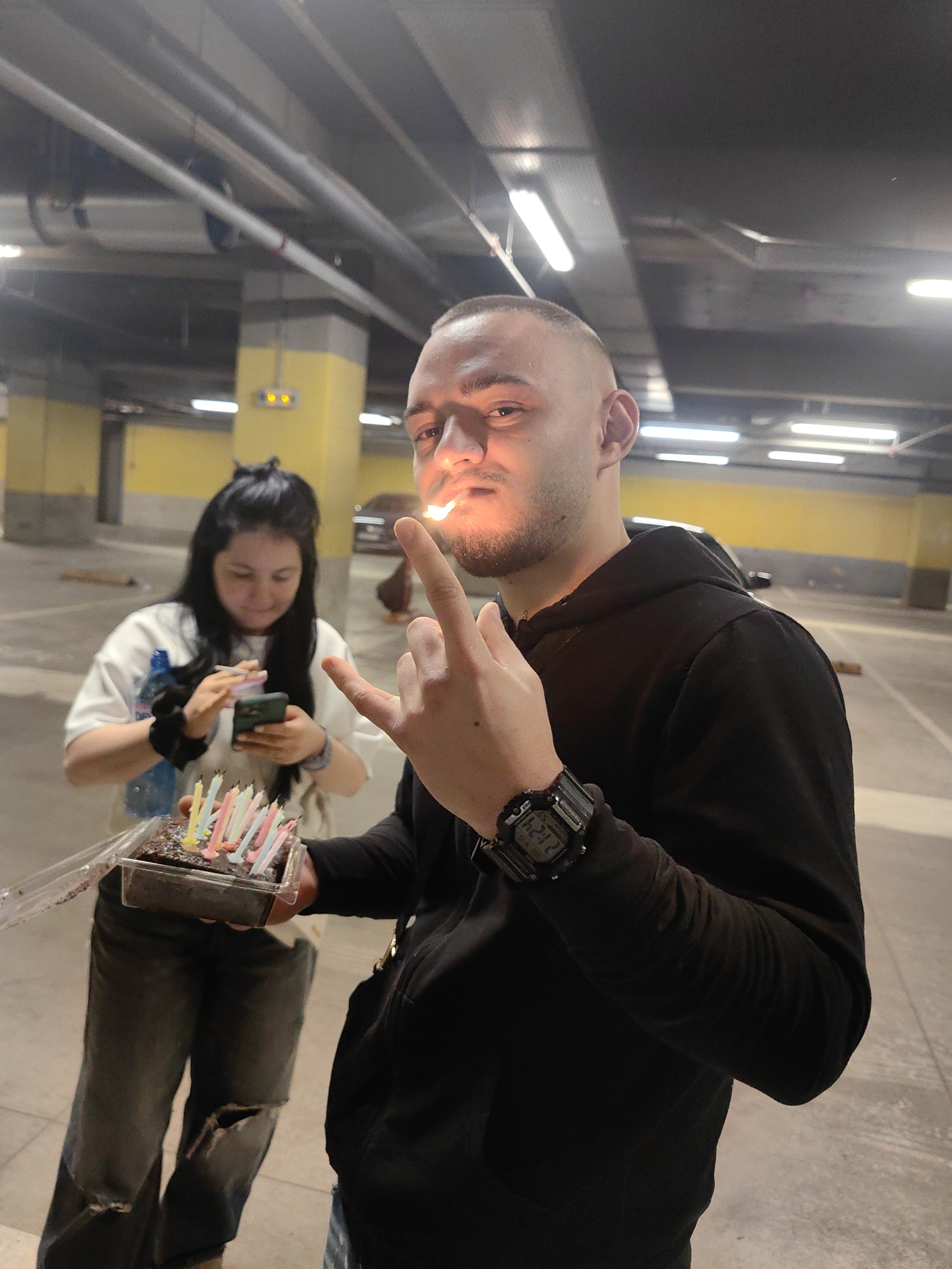 The image depicts a man holding a lit cigarette to his mouth, standing in what appears to be an underground parking garage. He is wearing a black hooded sweatshirt and a large digital watch. In his other hand, he holds a plastic container with a small, chocolate cake adorned with multiple lit birthday candles. A woman stands behind him, focused on her smartphone. The garage is concrete and dimly lit by fluorescent lights. The location cannot be specifically determined to be in Sofia, Bulgaria from just this image.