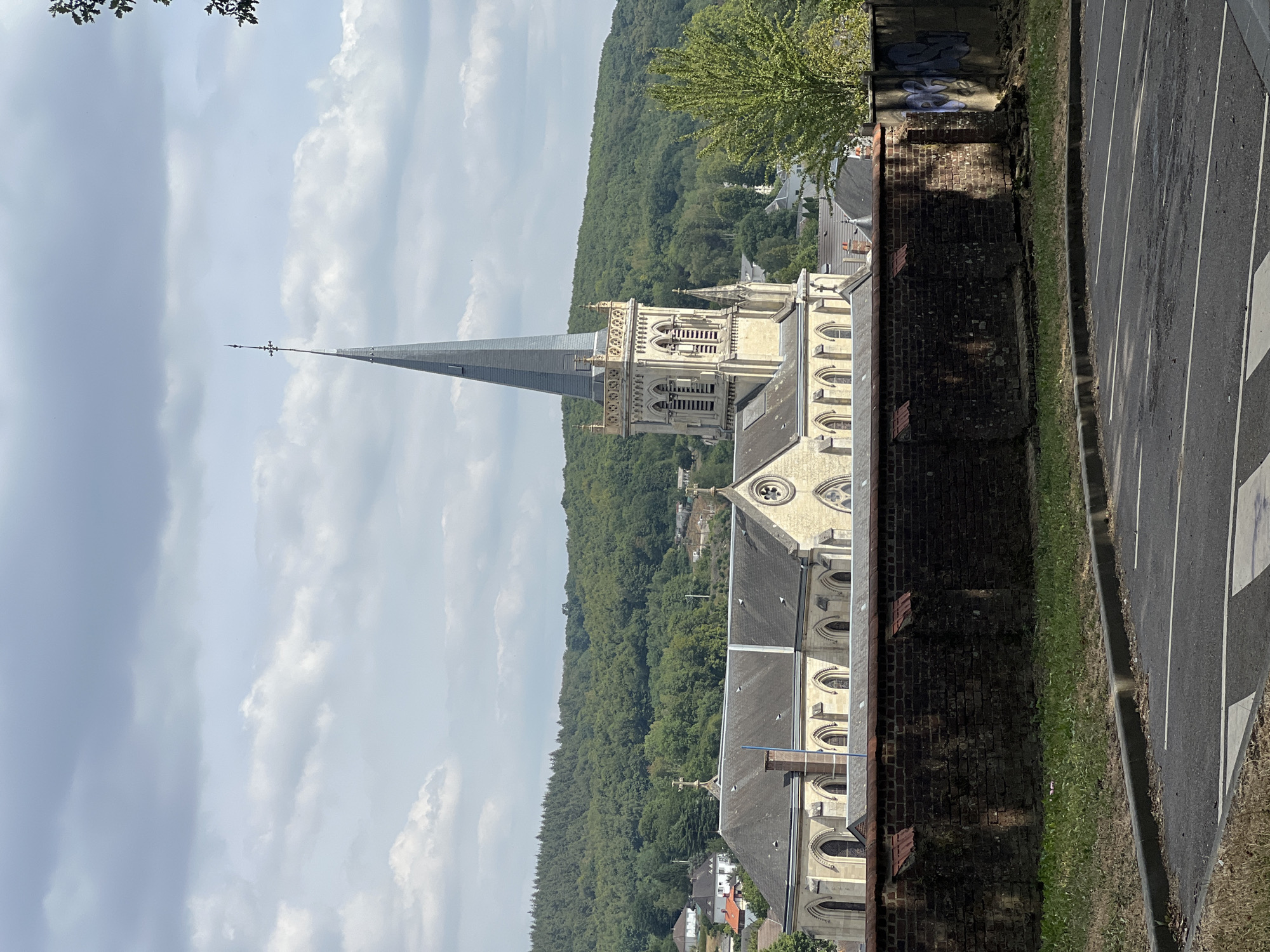 This image captures a view of the Église Saint-Martin in Le Houlme, France. The church, constructed from light-colored stone, features a prominent tall, grey spire topped with a cross. Its facade exhibits Gothic architectural elements, including arched windows and decorative stonework. The church is situated amidst a landscape of lush green trees and rolling hills, suggesting a semi-rural setting within or on the outskirts of the town. In the foreground, a low, weathered brick wall runs horizontally across the frame, partially obscuring the view of the church. Beyond the wall, a paved parking lot with white parking lines is visible. The sky above is partly cloudy, with patches of blue peeking through. The overall impression is one of a historic religious building set against a natural, verdant backdrop.