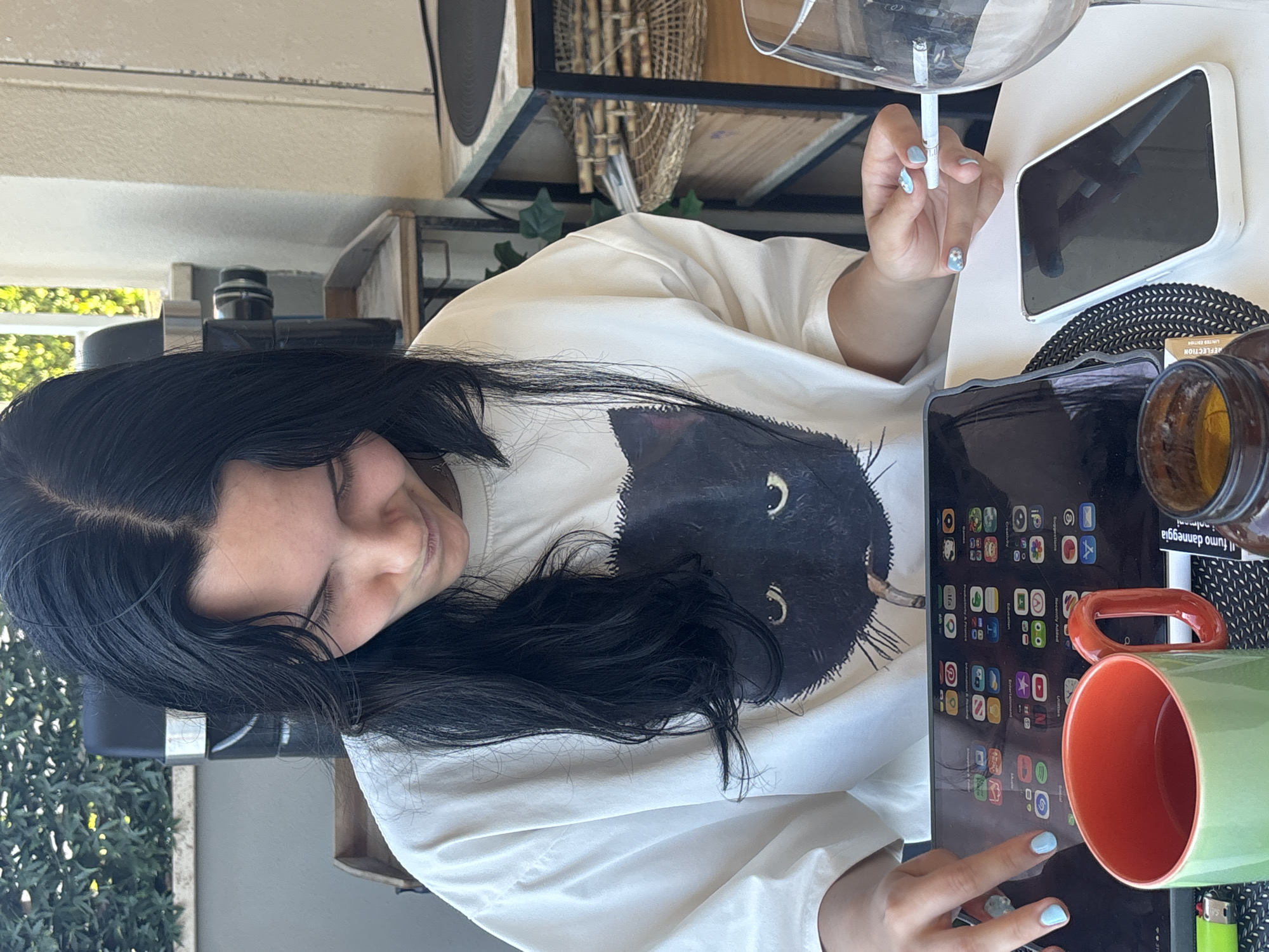 A young woman with long, dark hair is sitting at an outdoor table, looking down at a tablet computer. She is wearing a white t-shirt with a black cat graphic. Her fingernails are painted light blue and she is holding a cigarette in her right hand. On the table, in front of her, are the tablet, a green and orange mug, a smartphone, and a glass. A lighter is visible on the table next to the mug. In the background, there is a hedge, an outdoor grill, and shelves with various items. The scene appears to be on a balcony or patio in Cupra Marittima, Italy, given the visible text on a label that reads "FUMO DANNEGGIA LA SALUTE" which translates to "SMOKING DAMAGES HEALTH," a common warning in Italy.
