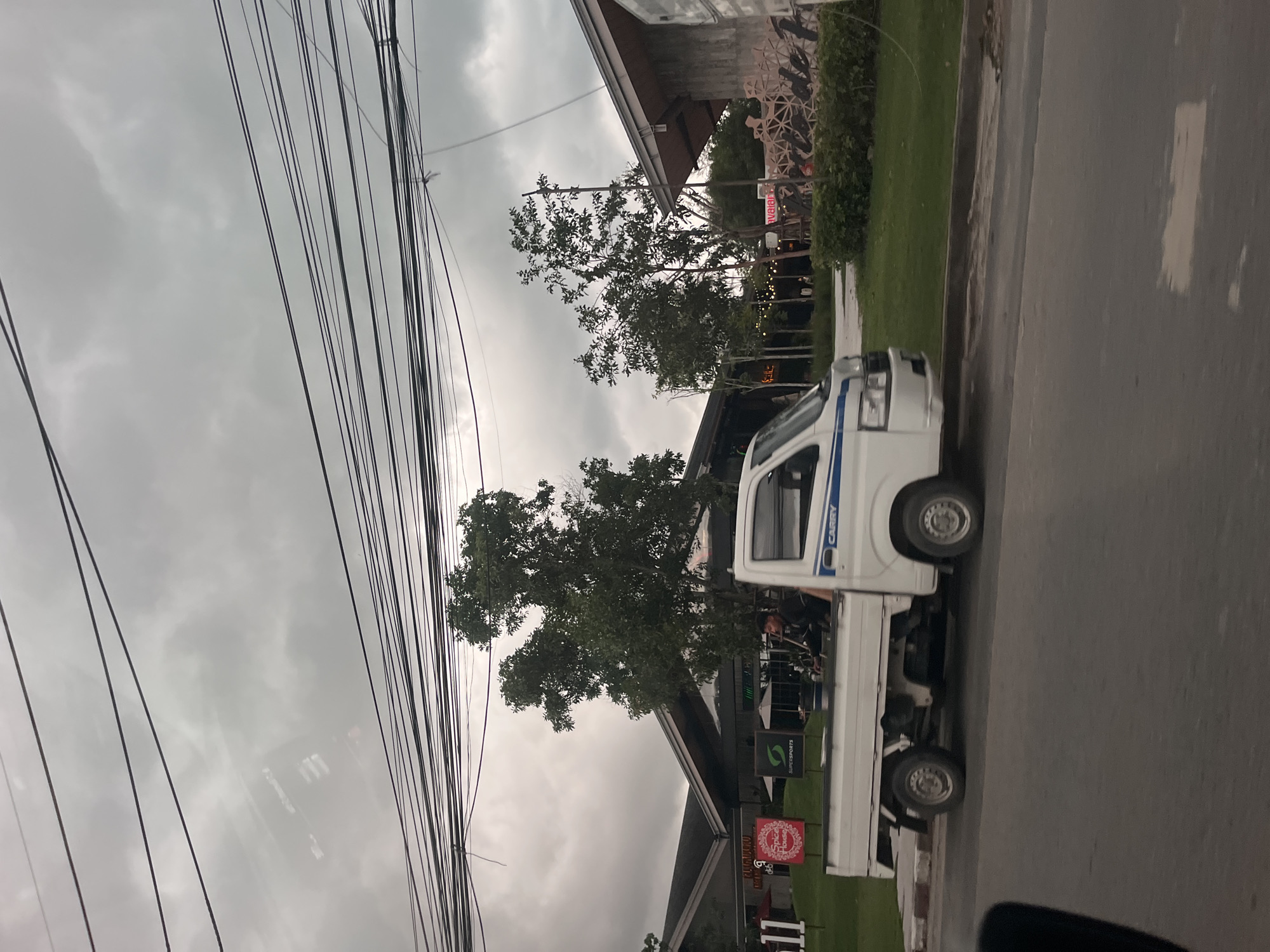 A white Suzuki Carry truck is parked on the side of a road in Choeng Thale, Thailand, under a cloudy sky. The truck is facing right, with its front wheels turned slightly towards the road. A person in a dark jacket is visible in the driver's seat of the truck. The truck bed appears empty. Behind the truck, a row of buildings with distinctive angled roofs are visible, some with signage indicating businesses. One sign reads "Spice House". Green grass and trees are situated between the road and the buildings. Power lines are strung across the top of the frame. The road in the foreground is asphalt and shows some discoloration.