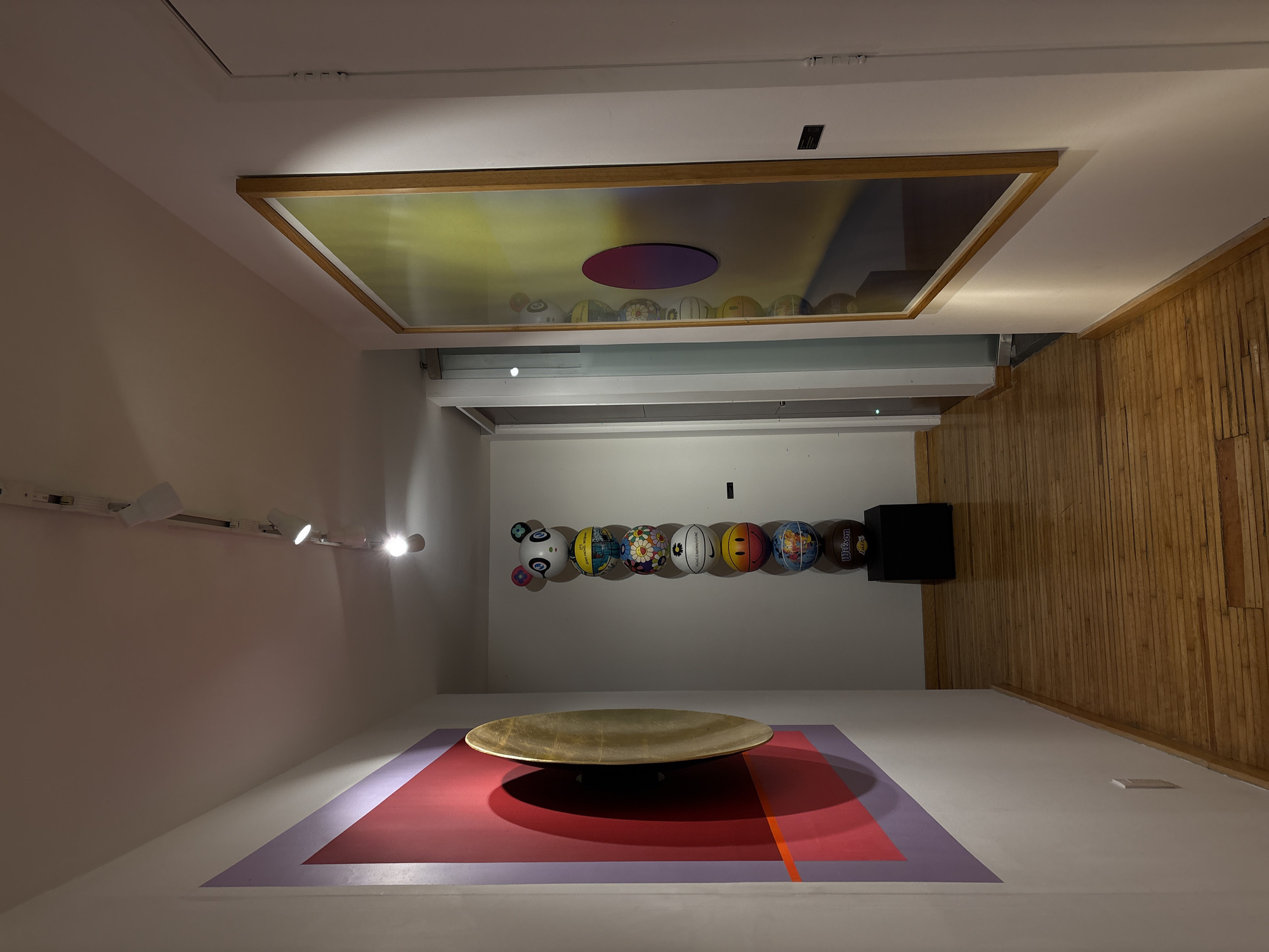 The image depicts a room in what appears to be an art gallery or museum, possibly located in Mexico City, Mexico, based on the artistic style and exhibition space. The perspective is from an elevated position, looking down.
The room features white walls on one side, contrasting with a wooden-paneled wall on the other. On the white wall, a track lighting system is visible. Overhead, there is a large framed art piece, containing a circular purple object with yellow and grey colors behind. Below the artwork and in line with it, there are a series of basketballs with different art drawn on each of them.
The center of the room features a large, oval-shaped, metallic-looking object (possibly a bowl or plate) placed on a rectangular surface of purple and red colors on the floor. There is also a bright orange line on the floor as well.