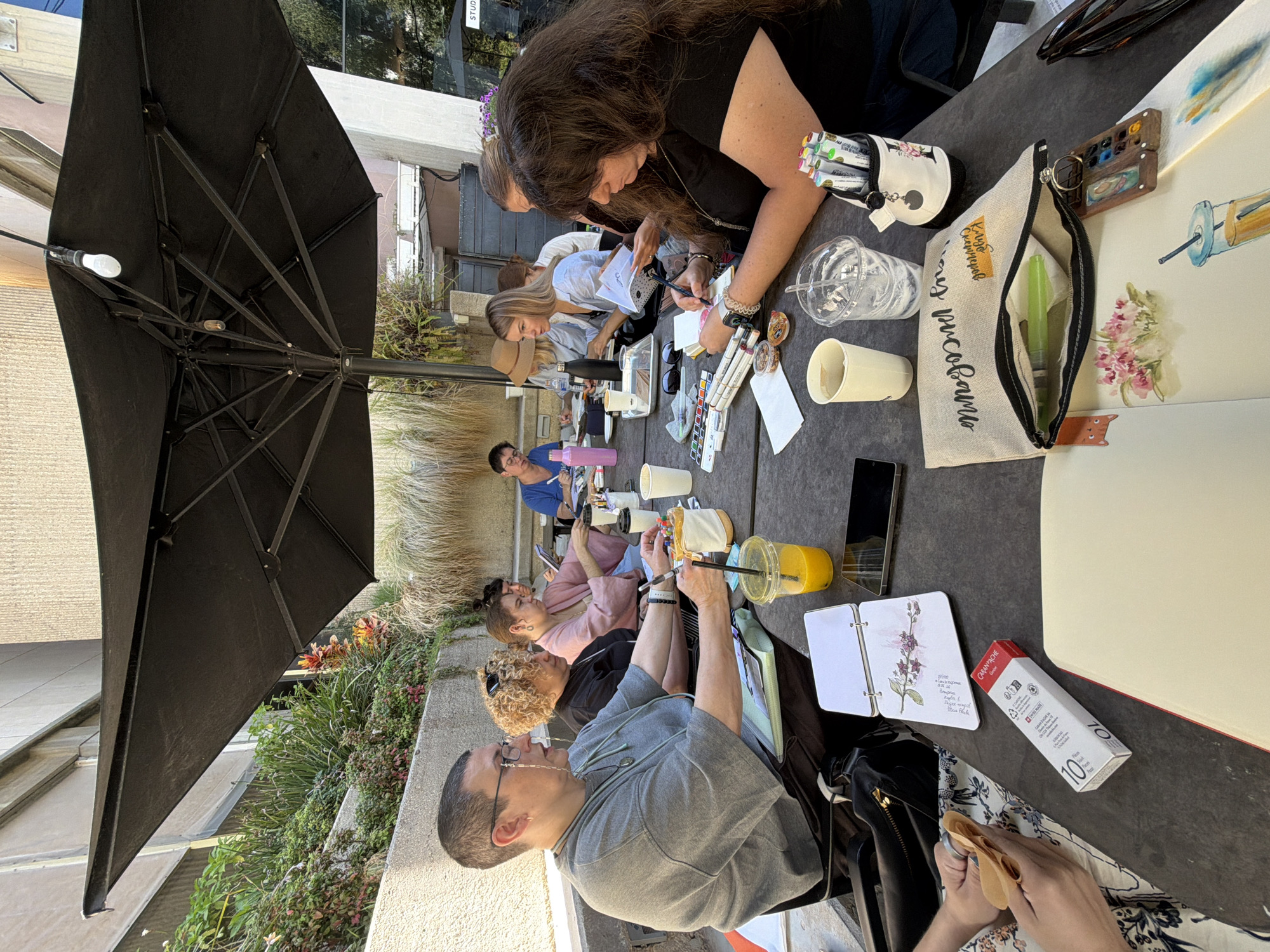 The image depicts an outdoor art class taking place on a patio, likely in Tel Aviv, Israel, given the architectural style and urban vegetation. A large black umbrella provides shade for a group of approximately seven individuals seated around a dark, textured table. The participants, a mix of genders and ages, are engaged in drawing and painting activities.
In the foreground, on the left side of the table, a person wearing glasses and a grey sweatshirt is seen from the side, with their hands holding a pen over a notebook. To their right, a woman with curly blonde hair, wearing a black top, is looking down at her artwork. Further to the right, a woman with long dark hair, wearing a black t-shirt, is actively drawing with a pen. Other individuals are visible in the background, also engrossed in their creative work.
The table is laden with art supplies, including sketchpads, notebooks with floral drawings, markers, pens, paint palettes, cups, and water bottles. One person is holding a stack of what appears to be origami paper. A black bag with an embroidered design is open on the table, revealing art supplies inside.
The environment suggests a bright, sunny day, with sunlight filtering through the umbrella. The background shows a textured wall, lush greenery, and hints of urban structures, including a building with large windows and signs, one of which reads "STUDIO". The overall scene conveys a relaxed and focused atmosphere of a creative workshop.