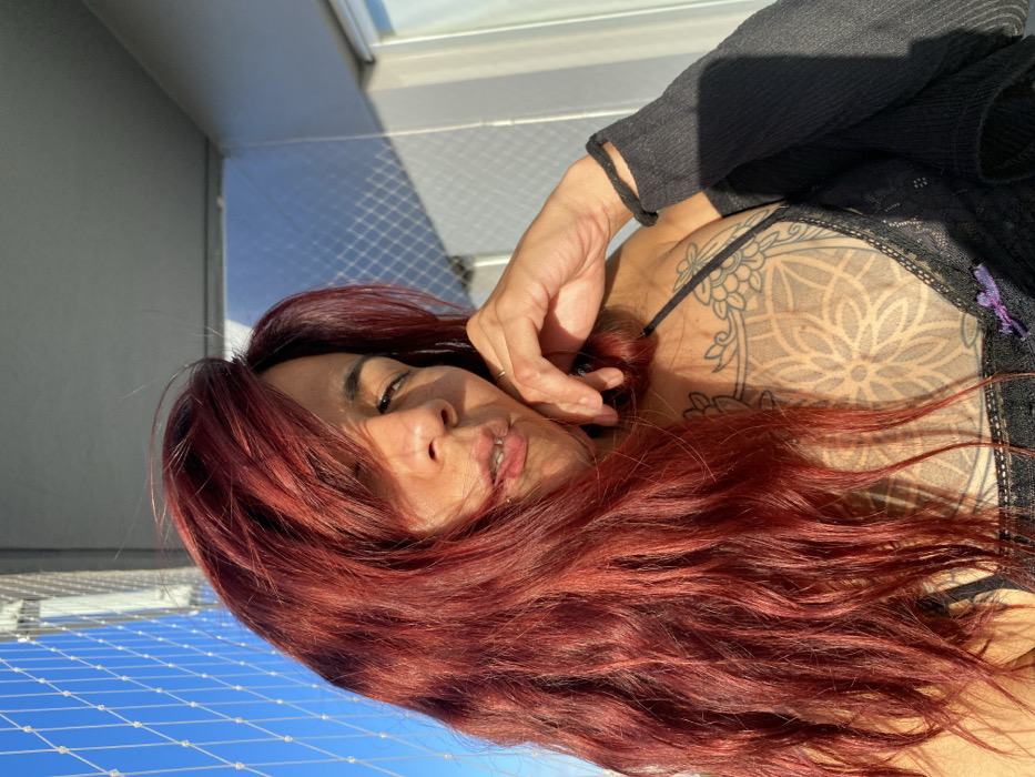 A woman lies on her side with fiery red hair cascading around her face. She wears a black top with lace detailing and a spaghetti strap underneath. A large, geometric tattoo is visible on her shoulder. Her hand rests near her face, fingers lightly touching her hair. The background appears to be a balcony with a net fence, revealing a bright blue sky. The photo is rotated, giving the impression of lying on a vertical surface. Sunlight casts shadows across her body and the background.