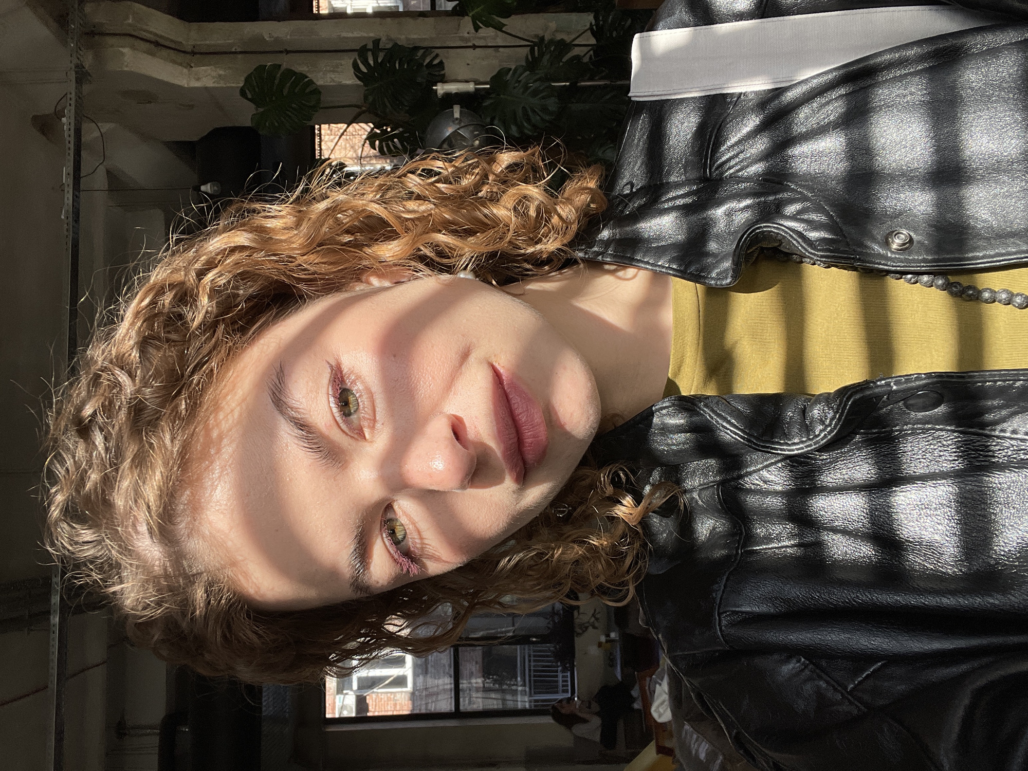 A medium close-up portrait shows a young woman looking directly at the camera. The image is oriented correctly for viewing, despite being presented sideways. She has fair skin, wavy, reddish-brown hair, and green eyes. Her expression is calm with a slight smile. She is wearing a black leather jacket over an olive-green top, with a white band or collar visible under the jacket. A dark beaded necklace is also visible around her neck.
The setting appears to be indoors, likely in a space with large windows, as evidenced by the strong, direct sunlight casting distinct, grid-like shadows across her face, jacket, and the background. These shadows suggest sunlight passing through a window with a grid pattern or blinds. In the blurred background, industrial-style elements like exposed pipes or ceiling structures are faintly visible, along with dark green, broad-leafed plants (resembling Monstera). Through a window in the background, distant buildings or structures can be vaguely discerned. The strong sunlight indicates a sunny day, likely morning or late afternoon.
The primary activity is posing for a portrait photograph. There is no visible text in the image. The location context provided is Tbilisi, Georgia.