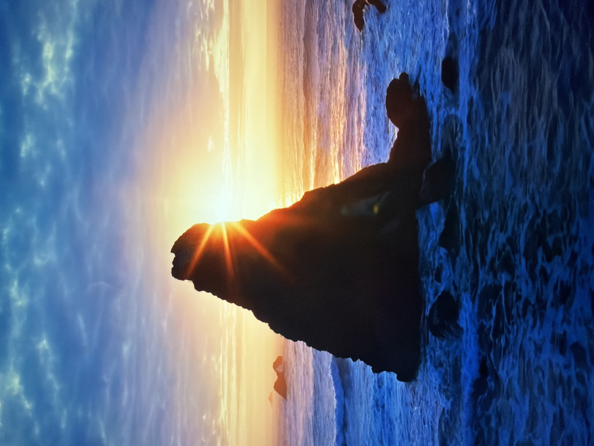 This is a vertical, mid-shot photograph taken at dusk. A large, dark, silhouetted rock formation dominates the foreground and left-center of the image. The sun is setting behind this rock, creating a bright lens flare and casting orange and yellow light across the sky and ocean. The ocean is depicted with active, deep blue waves crashing against the rock and surrounding smaller rocks. In the distance, the ocean stretches to the horizon where it meets a sky filled with wispy, multicolored clouds ranging from pale blue to orange. The scene is set by the sea, with rough, dark rock formations jutting out of the churning blue water. There are no people or man-made structures visible in the image. The location is not Kyiv, Ukraine.