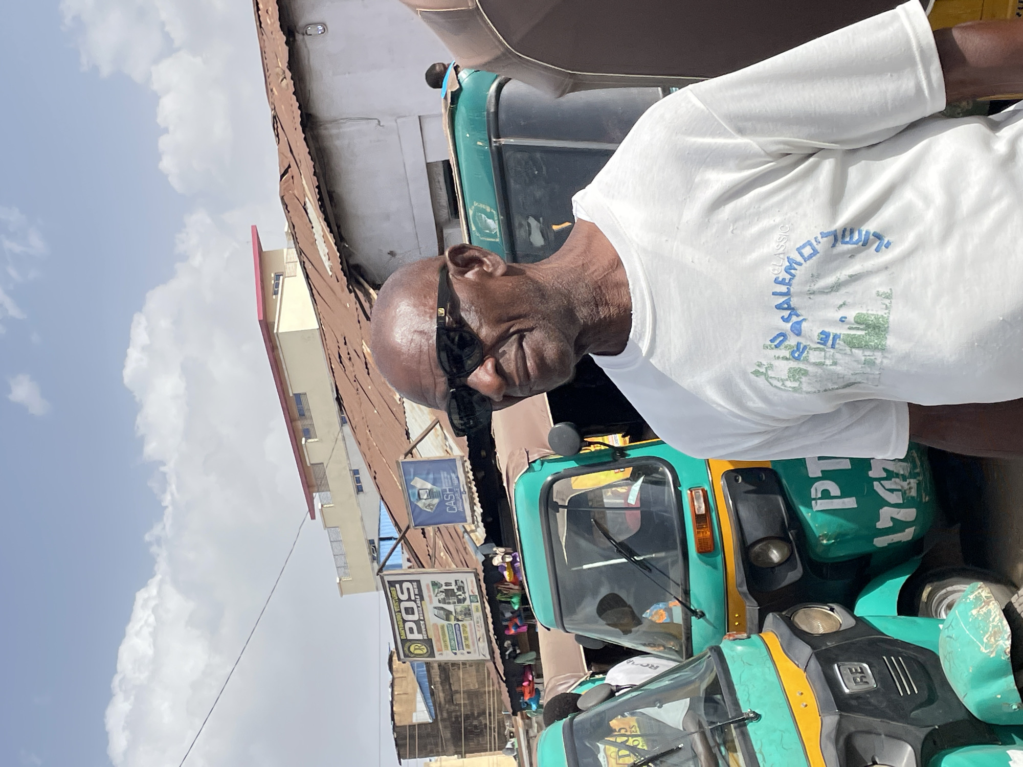 An outdoor, brightly lit image captures a street scene in Jos, Nigeria, featuring a smiling man and several tuk-tuks (auto-rickshaws) under a partly cloudy sky. The man, positioned in the foreground and slightly to the right, is seen from the chest up, looking towards the left with a subtle smile. He is a dark-skinned man with a bald head, wearing black sunglasses and a white short-sleeved t-shirt. His t-shirt bears a blue and green graphic design, with the English text "JERUSALEM CLASSIC" and Hebrew text "ירושלים" (Jerusalem) visible on the back.
The setting appears to be a busy urban or market area. Behind the man, multiple green and yellow tuk-tuks are parked or moving, indicating a common mode of transport. One tuk-tuk clearly displays the alphanumeric "PT 1747" on its side and "RE" on its front panel. In the background, various buildings with corrugated metal roofs and light-colored facades line the street. Several commercial signs are visible, including one advertising "CASH" services and another stating "DARYARORO OVERTURNED POS SERVICE AVAILABLE HERE", along with the partial text "ROA". The clear sky with scattered white clouds suggests a sunny day. The overall impression is a bustling snapshot of daily life in a Nigerian city.