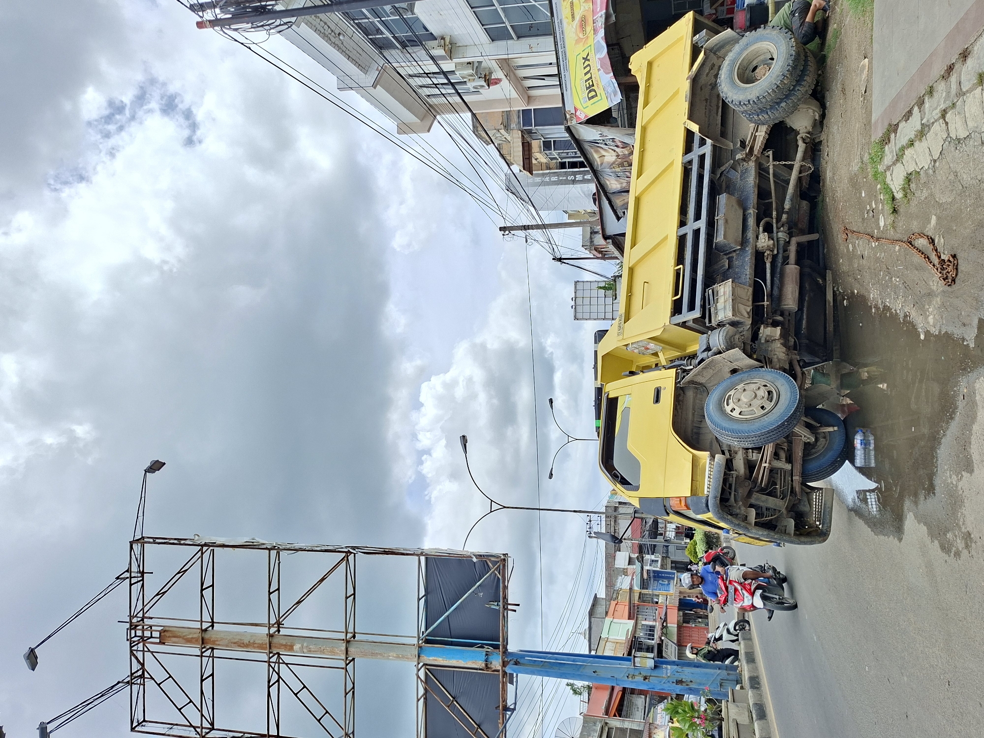 A yellow truck has overturned on a street in Lhokseumawe, Indonesia. The truck lies on its side, exposing its undercarriage and wheels. Nearby, three people on motorbikes appear to be observing the scene. A large billboard frame stands adjacent to the road. Buildings line the street and overcast skies provide general illumination. A chain lies on the sidewalk next to the overturned truck.