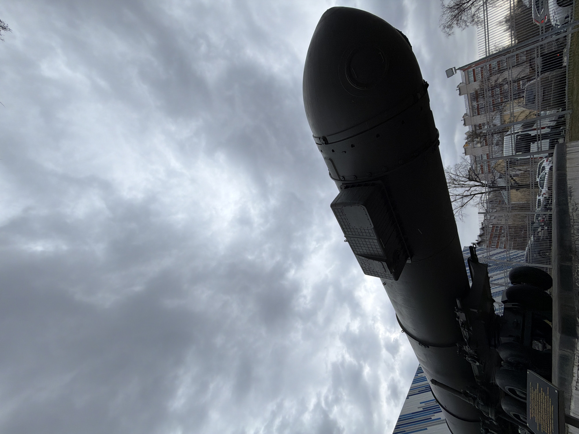The image presents a dramatic low-angle view of a large, dark-colored missile or rocket, silhouetted against a predominantly cloudy, overcast sky. The missile, likely a strategic ballistic missile, occupies the right half of the frame, with its conical nose cone pointing upwards. Visible details on its surface include seams, rivets, and box-like equipment housings. It rests on a large, multi-wheeled transporter, the dark chassis and robust wheels of which are partially visible at the bottom right.
In the background, behind a metal fence, are several multi-story buildings, appearing to be apartment blocks or industrial structures, with parked cars visible between them. Bare, deciduous trees suggest the season is late autumn or winter. A section of a modern building with a distinctive blue and white striped or paneled facade is visible at the bottom left, possibly part of a museum or public facility.
The heavy, gray cloud cover indicates an overcast day. The missile's static display, along with what appears to be an informational plaque (text illegible) near the transporter's wheels, suggests it is an exhibit or memorial. Given the location context of Votkinsk, Russia, known for its machine-building plant that produced ballistic missiles, this is likely an RT-2PM2 Topol-M (or similar) missile on display, commemorating the region's contribution to defense and industry. The scene evokes a sense of historical significance and solemn power.