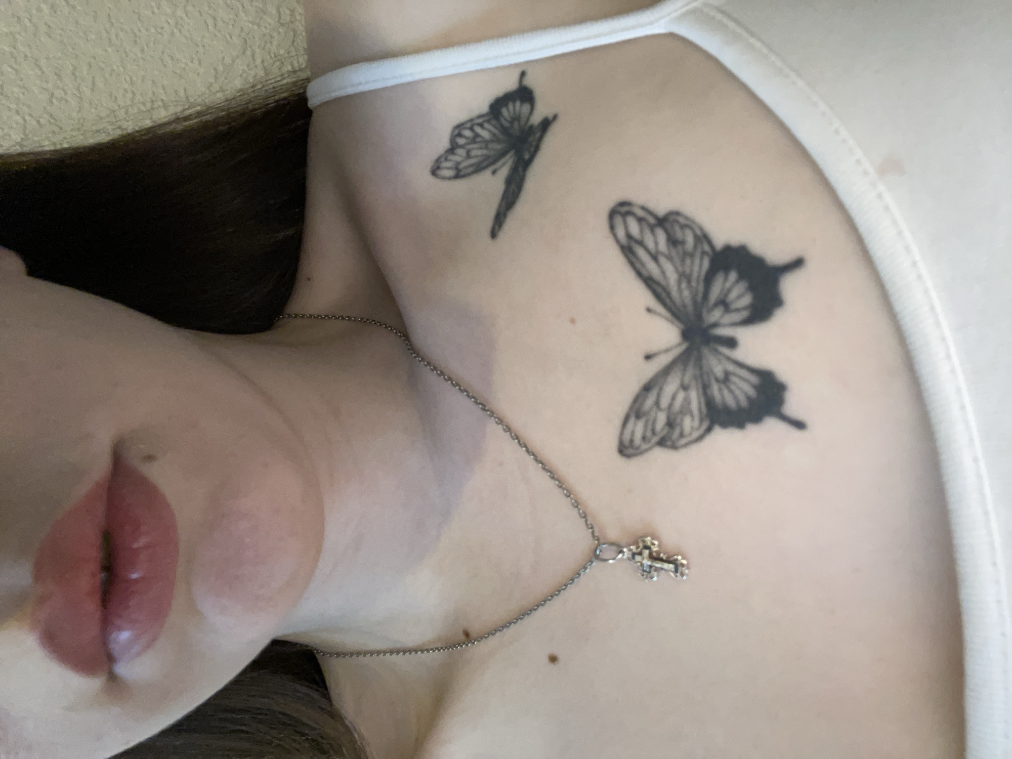 A close-up, angled shot of a person's upper chest and face. Two butterfly tattoos are visible on the upper chest and shoulder. A silver necklace with a small cross pendant rests on the chest. The person has dark hair and pink lips. The background appears to be a plain wall. It is not possible to determine the location in Chaikovsky, Russia from the given image.