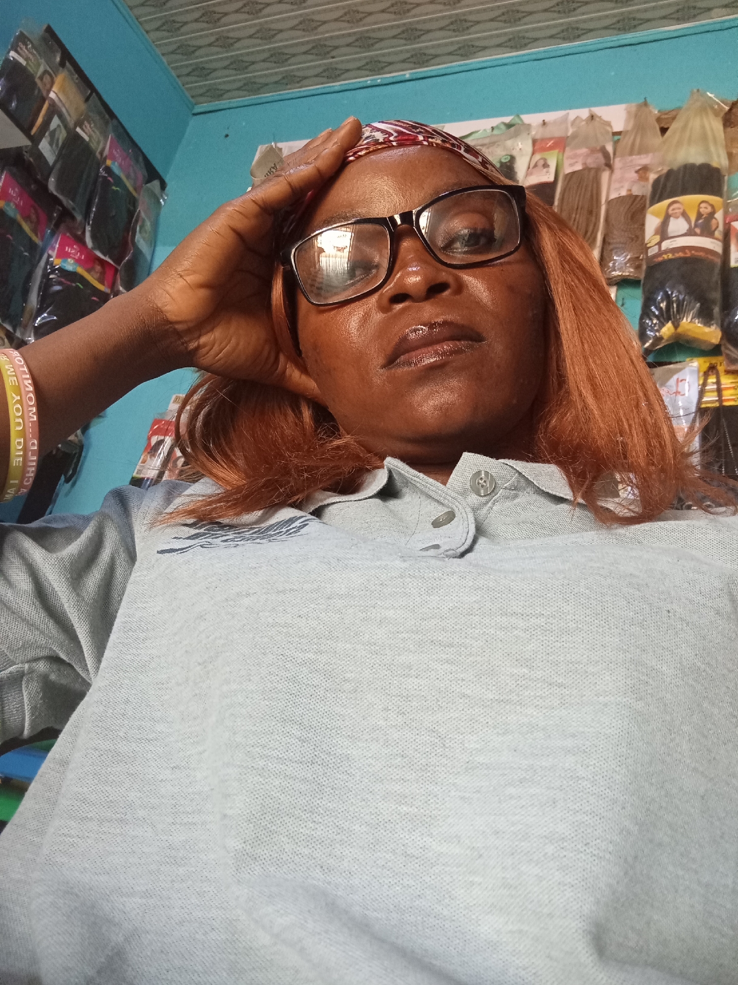An overhead, medium shot captures a woman wearing glasses, a patterned headscarf, and a grey polo shirt. She has medium-length, reddish-brown hair. Her right hand is raised to her forehead. Behind her, shelves are stocked with numerous packaged hair extensions. The wall behind the shelves is painted light blue, and the ceiling is textured and white. The overall scene suggests an indoor retail environment, possibly a hair salon or a shop selling beauty products, located in Jalingo, Nigeria.