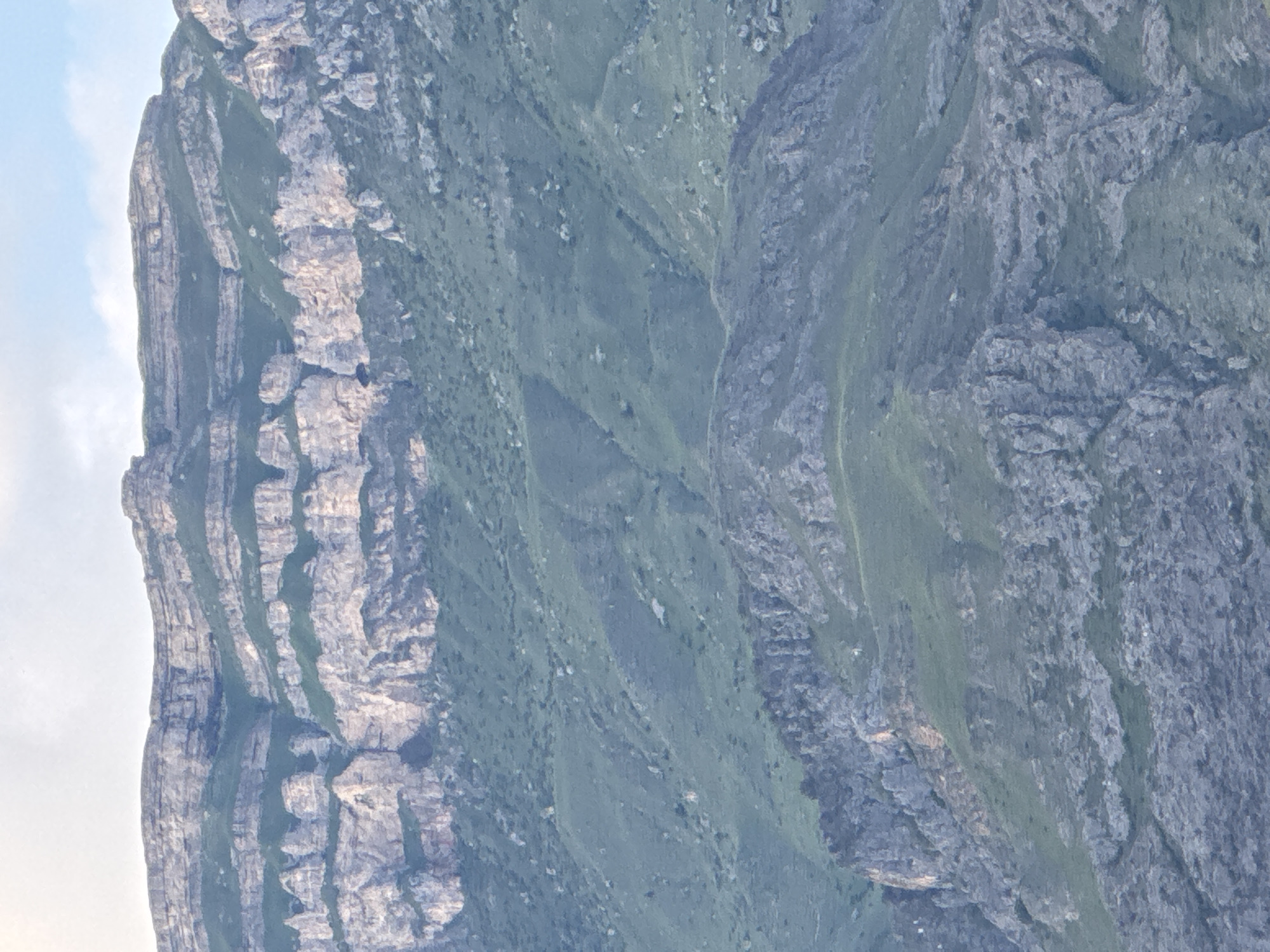 The image displays a panoramic view of mountainous terrain with stratified rock formations and green vegetation. The foreground features rocky slopes interspersed with patches of grass, while the midground shows more rounded, vegetated hillsides. The background is dominated by a large mountain range with distinct horizontal layers of rock, varying in color from light tan to gray, and streaks of green vegetation growing in the crevices. The sky above is a pale blue with some white clouds. The scene appears to be a natural landscape, likely within the region of Khadzhalmakhi, Russia, known for its mountainous geography. There are no people or man-made objects visible in the image, suggesting a remote or natural setting.