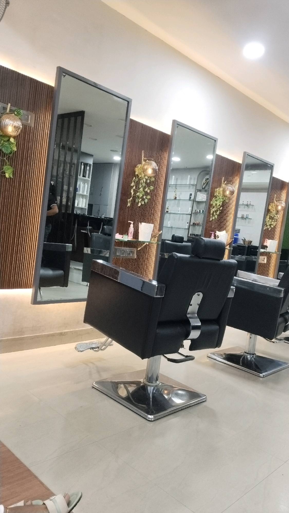 The image shows a row of styling chairs and mirrors in a beauty salon. The chairs are black with silver accents and hydraulic bases. The mirrors are tall and framed, with wooden paneling behind them. Decorative hanging plants and globe lights are mounted on the wooden panels. The floor is tiled in a light beige color. In the background, shelves stocked with various products are visible. One mirror reflects a person standing, presumably a stylist or customer, in a black shirt. The salon appears to be modern and well-lit.