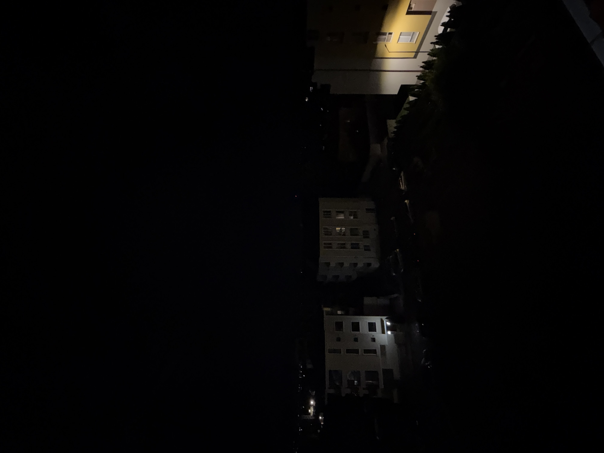 The media file presents a high-angle, nighttime view of an urban environment in Plovdiv, Bulgaria. The image is significantly dark, with the entire left half appearing as an expanse of deep black, possibly indicating a shadow, an unlit area, or part of a structure from which the photo was taken.
On the right side, a vertical strip reveals several multi-story buildings and a line of dark foliage. A prominent building at the upper right is light yellow with white-framed windows, some of which are illuminated. Below it, other structures are visible, appearing white or light gray, with numerous windows. Several of these windows also show interior lights, indicating active spaces within the buildings. A dense line of trees or bushes runs along the edge of some buildings, appearing dark against the faintly lit structures. Faint points of light are discernible at what seems to be ground level towards the bottom right, possibly from distant streetlights or vehicle lights.
There are no people visible, and no specific activities are taking place, creating a static and quiet atmosphere. The time of day is clearly night, with all visible illumination originating from artificial light sources within the buildings or from distant street lighting. The perspective suggests the photograph was captured from an elevated position, looking down into a dense urban or suburban area. No visible text is present in the image.