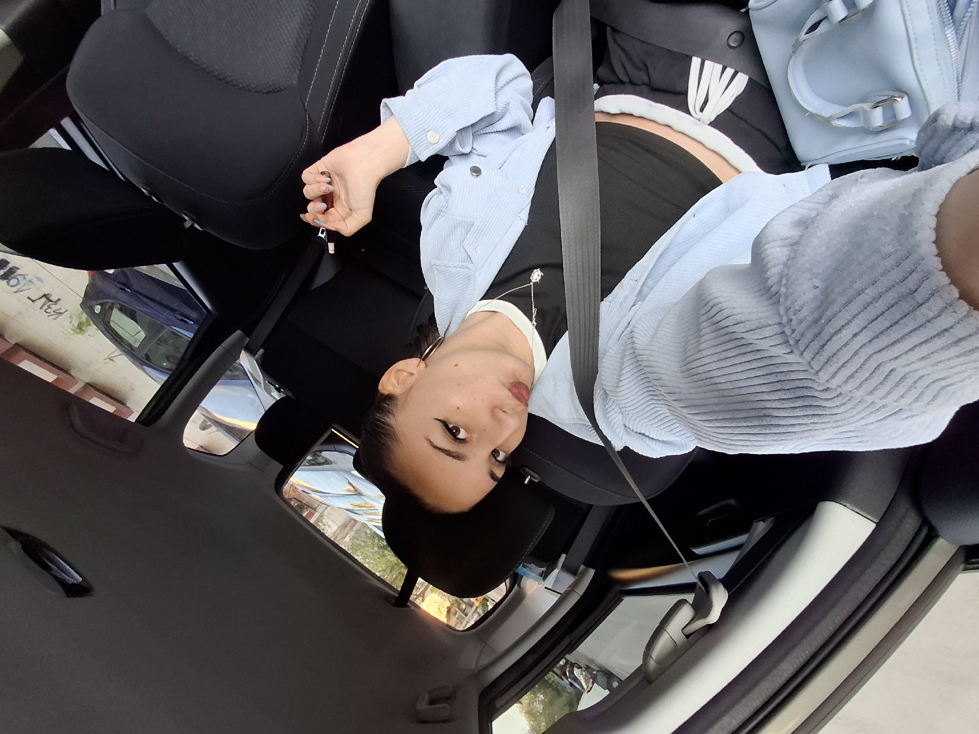 A young woman is captured in a selfie or photo taken from within a car, likely in Sofia, Bulgaria, during the daytime. She is positioned in the front passenger seat, wearing a seatbelt, and looking directly at the camera with a slightly pouted expression. Her dark hair is pulled back, and she has prominent winged eyeliner and dark lip makeup.
She is dressed in a light blue, ribbed long-sleeved shirt, possibly worn open over a black crop top or bandeau that exposes a sliver of her midriff. A delicate necklace with a star-like pendant adorns her neck. Her right hand, with manicured nails featuring light blue tips, holds a red-brown pen or vape device.
The car's interior features black seats and a grey ceiling and trim. A light blue structured handbag rests on the seat beside her, and a dark jacket or hoodie with white striped detailing is visible behind her. Through the car windows, an urban or suburban environment is visible, including other parked vehicles (one dark blue car is distinct), buildings, and what appears to be a street or sidewalk with some faint, unreadable graffiti. The natural light suggests a clear day. The overall impression is a casual, posed moment inside a vehicle.
