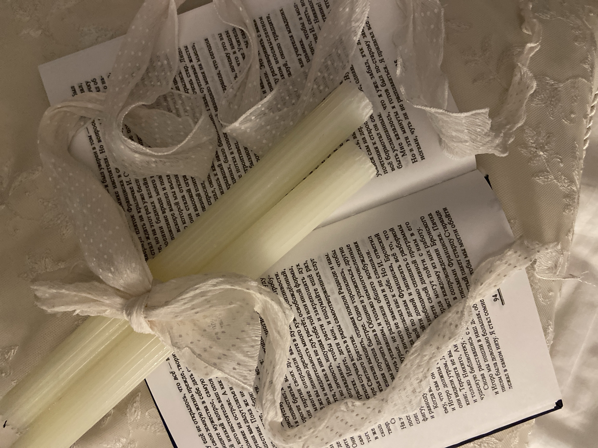 Here's a detailed description of the image:
**Scene:** A photograph shows two off-white candles tied together with a white ribbon adorned with small dots, resting on an open book. The book lies on a cream-colored, lace-textured surface, suggesting a soft, delicate setting.
**Objects:**
* **Candles:** Two cylindrical, off-white candles tied in the center with a ribbon.
* **Ribbon:** The ribbon is white with small dots and forms a bow around the candles.
* **Book:** An open book with printed text in a language using Cyrillic script. The pages are slightly aged, with a warm tone.
* **Lace Surface:** A cream-colored, lace-like fabric providing the background and base for the objects.
**Text (based on OCR):**
The text on the book pages includes partial phrases and words like:
"еще не открылось, что все - в", "свою земную жизнь прежде, твори", "ищите мужества в себе дать г", "домике настоятеля, но вопрос...",
There are also phrases such as "...что в нашем...", "...наводило меня на мысль", and "...что сегодня".
There is a page number "94" visible.
**Environment:** The environment appears to be indoors, with an emphasis on creating a soft, aesthetic, and literary atmosphere. The lighting highlights the textures and creates a gentle mood.
**People, Subjects, Actions, Interactions:**
While people are not explicitly visible, the presence of the candles, ribbon, and book suggests human arrangement and intention. The book's content implies intellectual or spiritual interests.
**Location:**
Given the use of the Cyrillic script and the information on the text of the book, it is likely this image does not belong to the city of Budva, Montenegro, since the official language is Montenegrin, and the main language is not using Cyrillic script in everyday communications.