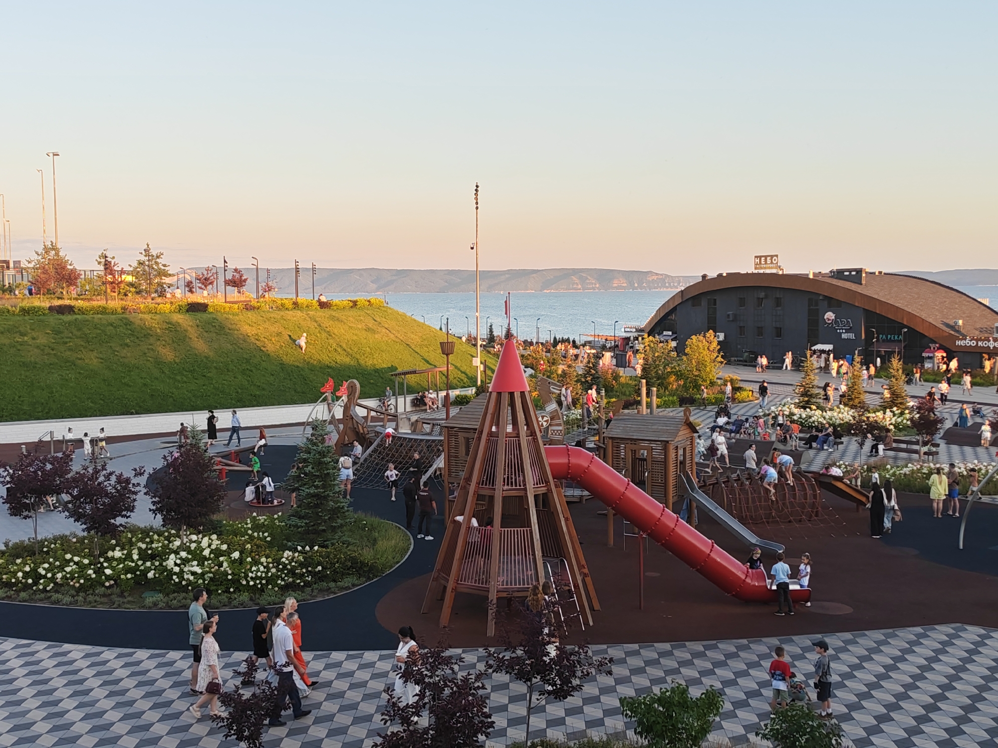 This is a high-angle, wide shot of a modern, outdoor public space, likely a park or waterfront promenade, bathed in the warm, golden light of late afternoon or early evening. The scene depicts a recreational area with a playground prominently featured in the foreground and midground. The playground includes a tall, wooden conical structure with a red slide, climbing nets, and smaller wooden playhouses. Children and adults are visible interacting with the play structures, with some children playing and others observing.
To the left of the playground, a grassy, sloped embankment is covered with landscaping, featuring trees and flowering bushes, interspersed with lampposts. Beyond this, a body of water, likely the Volga River, stretches across the horizon, with distant hills or mountains visible on the opposite bank.
In the background, a large, dark, curved building with a brown roof is situated. Signage on the building includes "NEBO" (Russian for "sky") and "TASTEOPPORTUNITY" in English, as well as "APADA HOTEL" and "REKA" (Russian for "river"). People are seen walking along pathways and gathered around the building. The ground in the foreground and midground is paved with a distinctive black and white checkered pattern.
The overall impression is one of a well-maintained, popular urban recreational space designed for leisure and family activities, with a scenic backdrop of a river and distant landscape. This location is likely the Tolyatti embankment, a popular recreational area in Tolyatti, Russia.