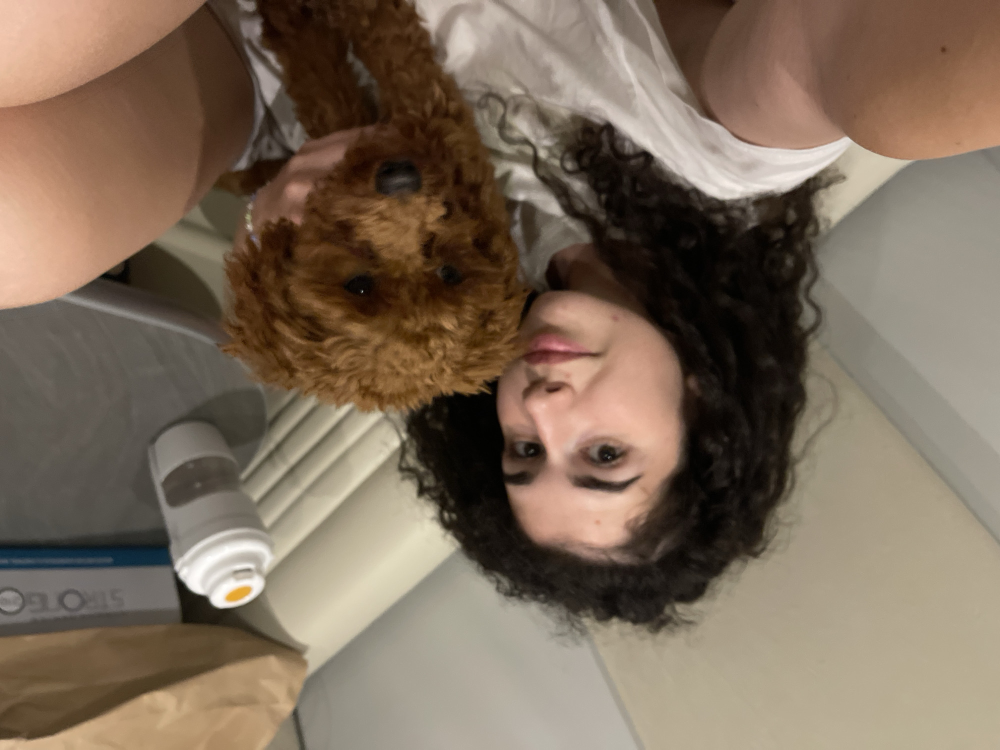 The image captures an indoor scene from an elevated, vertical perspective. A young woman with dark, curly hair is positioned on her back, looking towards the camera. She has defined eyebrows and appears to be wearing a light-colored lip product. Her torso and upper arm, covered by a white top, are visible. Adjacent to the woman, a small, reddish-brown dog with curly fur, resembling a Poodle, is centrally featured. The dog's face and dark eyes are oriented towards the camera. One of the woman's hands is gently placed near the dog's neck or head. Both the woman and the dog are situated on a light, neutral-toned surface, likely a bed or couch. In the upper left portion of the background, a brown paper bag stands next to a white rectangular box, partially revealing the text "STROTEC". A white, cylindrical container with an orange cap lies horizontally near these items. A light-colored wall or partition forms the rear background of the scene. This scene is located in the city of Koprinka, Bulgaria.