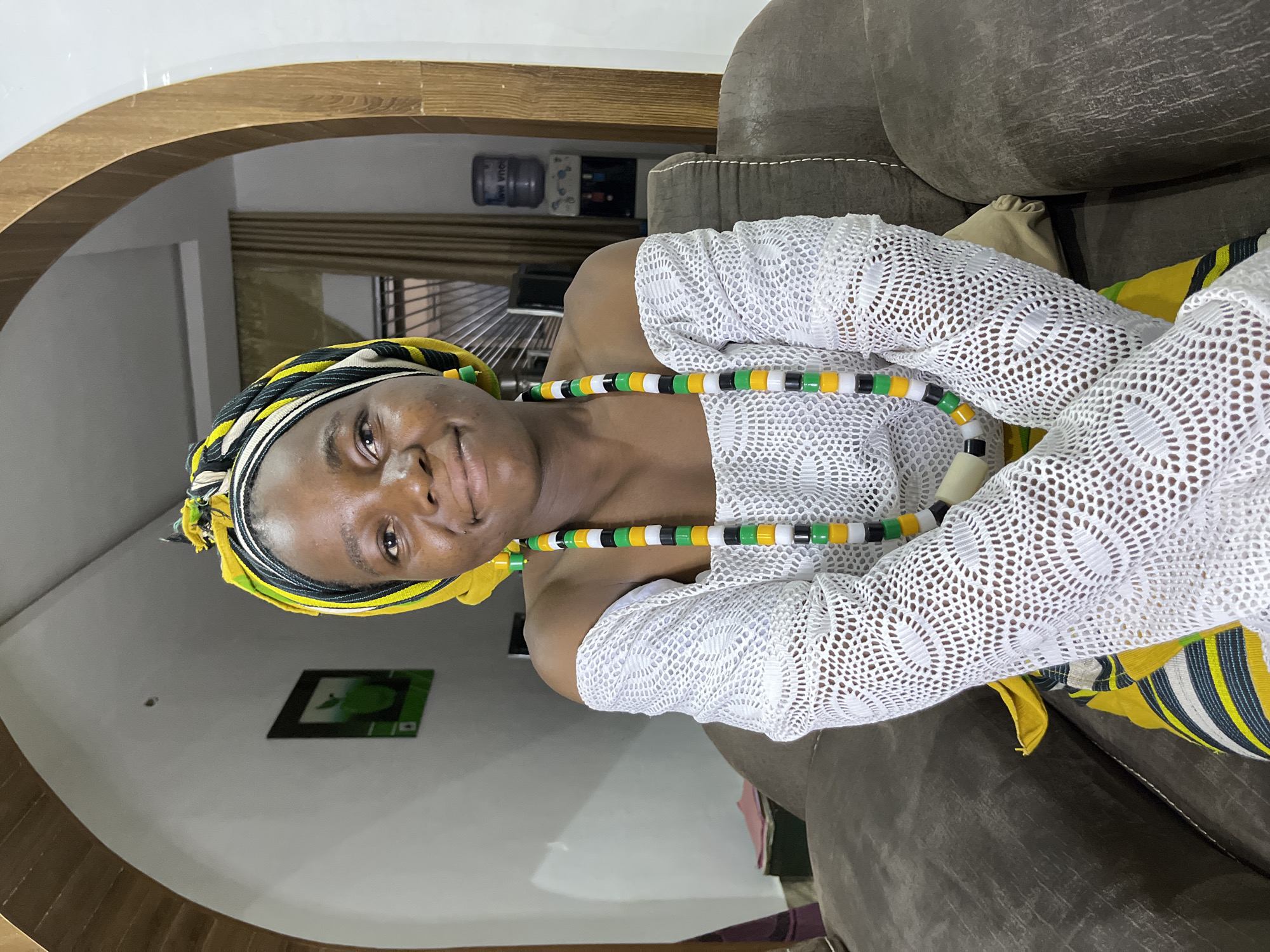 A woman is indoors in Jos, Nigeria. She wears a yellow and black headscarf, a white lace top, and a necklace with green, black, white, and yellow beads. She is seated on a brown couch under a wooden archway. A framed picture hangs on the wall. A water dispenser is visible in the background.