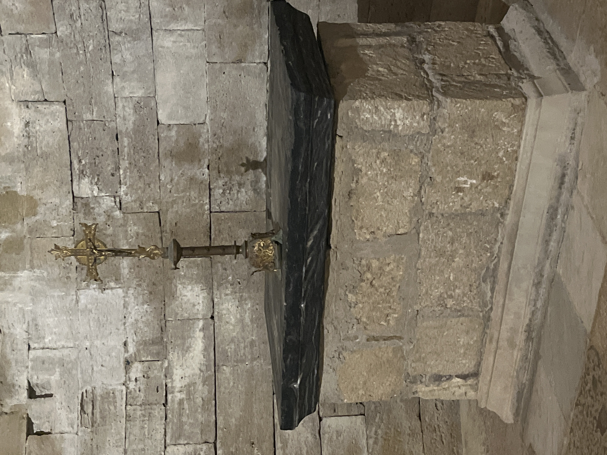 The image shows a close-up, eye-level view of a small altar or shrine within a stone structure. The background consists of rough-hewn, light-colored stone blocks arranged in horizontal courses, with visible mortar lines. Dominating the foreground is a dark, rectangular stone slab resting on a base made of larger, rougher stone blocks. On top of the dark slab, a golden crucifix with an ornate base stands prominently, casting a faint shadow on the stone behind it. The overall impression is one of antiquity and solemnity, likely located in a historical or religious site. This altar is located in the crypt of the Cathédrale Saint-Jean-Baptiste de Maguelone in Villeneuve-lès-Maguelone, France.