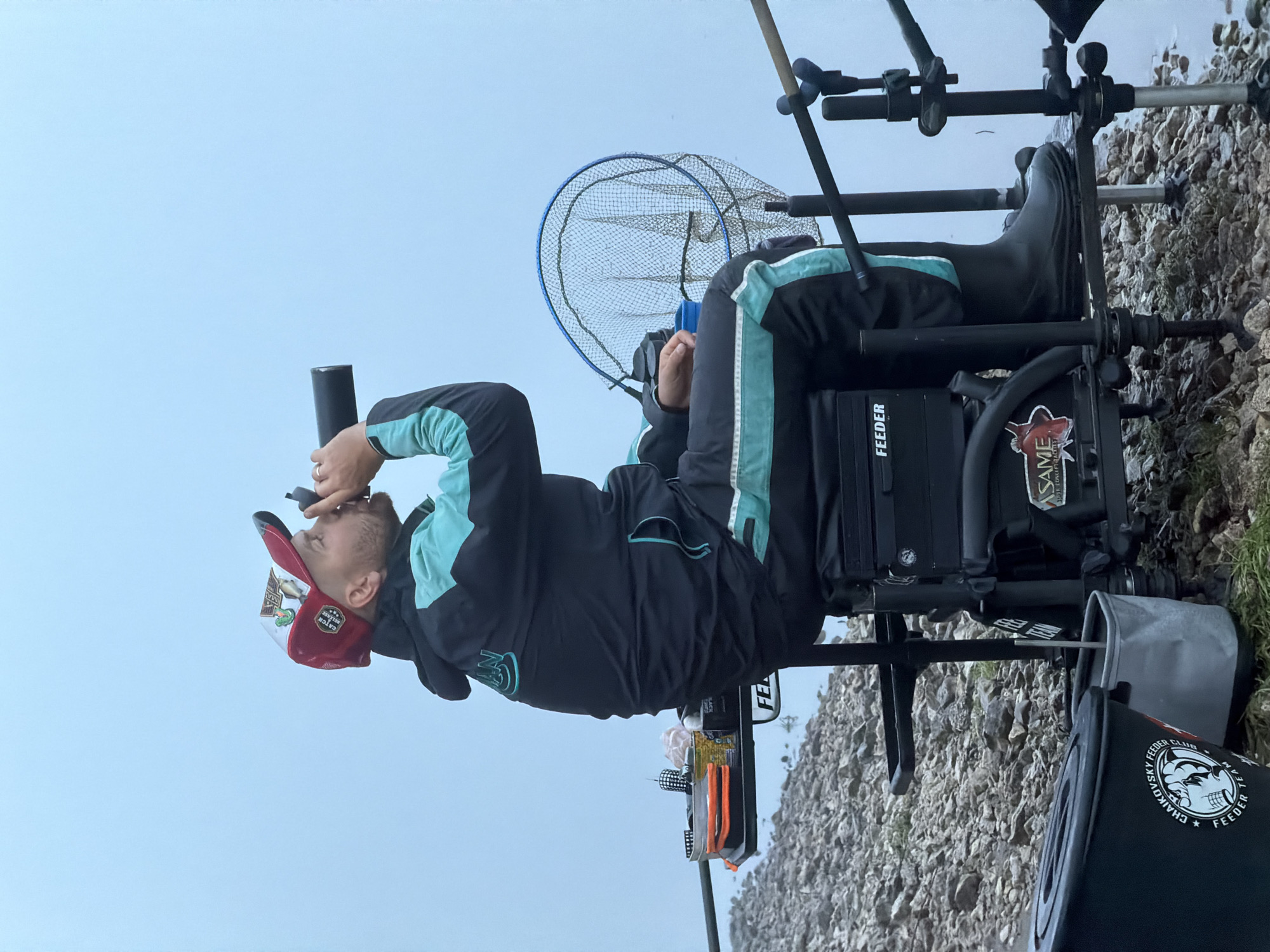 An angler is seated on a specialized black fishing platform at the edge of a body of water, likely a river or lake, on a rocky or gravelly bank. The image is taken under an overcast or pale blue sky, suggesting daytime with diffused light.
The man, wearing a dark jacket with light blue accents, black rubber boots, and a red and white baseball cap, holds a black cylindrical device (possibly a monocular or rangefinder) to his right eye, appearing highly focused on the water. His fishing setup includes multiple rods mounted in front of him, and a blue-rimmed landing net is visible behind his seat. Several black buckets and containers are positioned around the fishing chair.
Visible text on his gear includes "FEEDER" and "ASAME SURF CONCEPT" on the fishing seat, with an "ASAME" fish logo. His cap displays "CATCH FEEDER." A black bucket prominently features "CHAIKOVSKY FEEDER CLUB" and "FEEDER TEAM" along with a distinctive skull logo, indicating a specific fishing organization. The scene depicts a dedicated fishing activity, likely feeder fishing, in Chaikovsky, Russia.