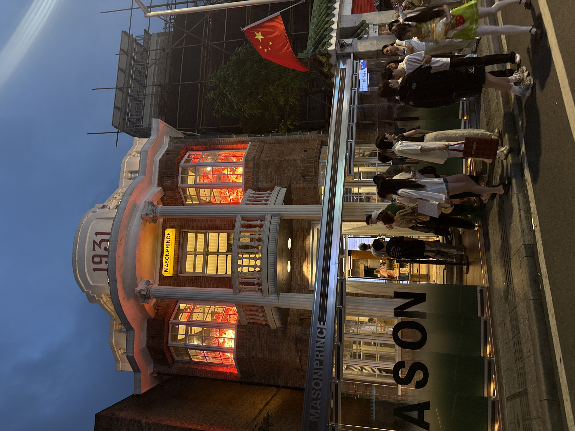The image shows the Mason Prince building, built in 1931, in Haizhu District, China. The building is three stories with brickwork, white columns, and lit windows. A sign with "MASON PRINCE" is visible on the building. The Chinese flag is shown flying above the building. There are people walking on the street. Scaffolding is visible on the top of an adjacent building.