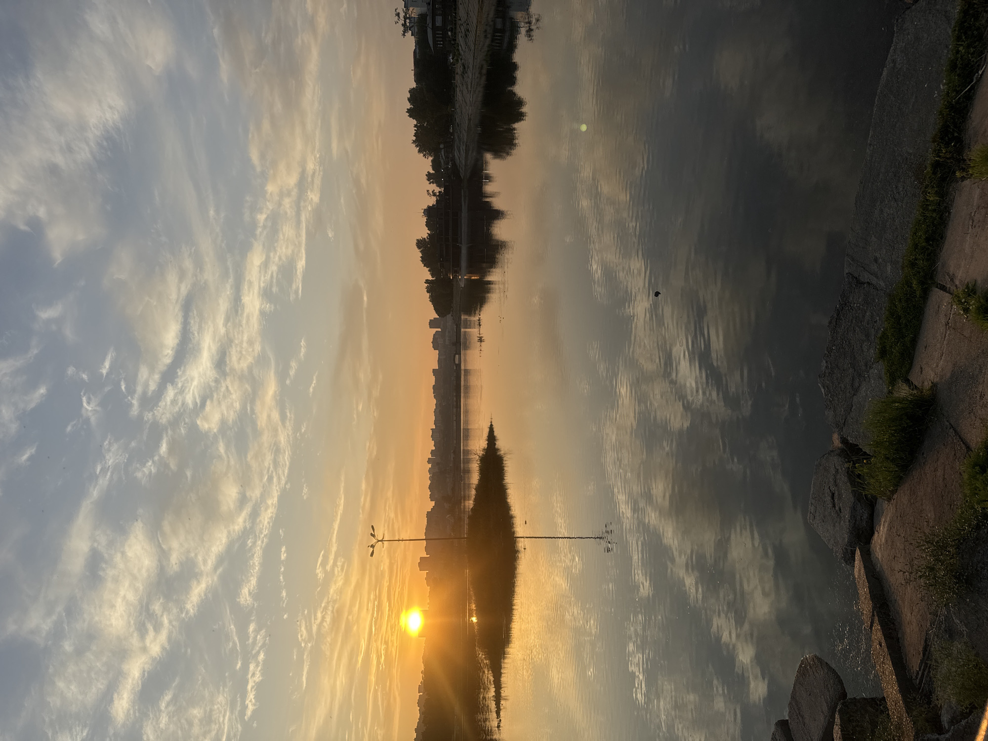 This is a vertical, medium shot photograph of a sunrise over a body of water, likely a river or lake, in Irkutsk, Russia. The sun, a bright, intense orb, is low on the horizon, casting long, golden rays across the scene. It is partially obscured by wispy clouds in the sky, which are painted in shades of orange, yellow, and blue. The clouds are scattered and thin, allowing the light to break through.
In the foreground, large, rough-hewn stones line the edge of the water, with patches of green grass growing between them. The water itself is calm and reflective, mirroring the sky and the distant cityscape. A small, grassy island with a single lamppost stands in the middle of the water, its reflection clearly visible.
In the mid-ground, the shoreline is visible, featuring a line of trees and some buildings, including what appears to be a multi-story structure with a dark roof, possibly a restaurant or boathouse. Further in the distance, a cluster of modern buildings forms the cityscape.
The overall mood of the image is serene and peaceful, capturing the quiet beauty of a sunrise in an urban environment. No people are visible in the frame. The focus is on the natural elements and their interplay with the man-made structures.