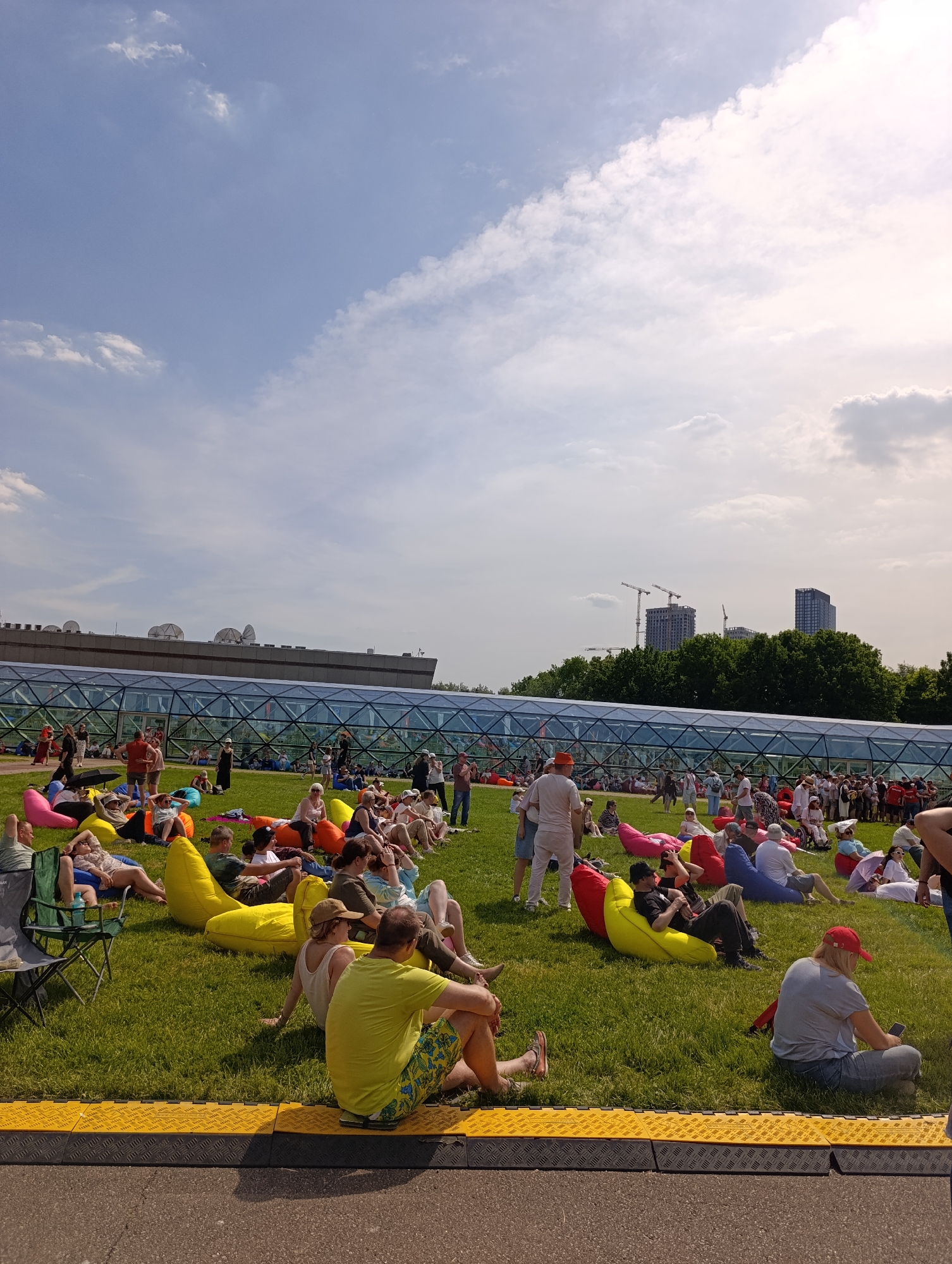 The image displays an outdoor scene in Zaryadye Park, Moscow, Russia, under a bright, partly cloudy sky. The foreground features an asphalt path bordered by a yellow and black textured rubber matting system. Beyond this, a wide, open expanse of green grass is populated by numerous people.
Approximately 50 to 70 individuals are visible, primarily seated or reclining on an assortment of colorful bean bags in shades of yellow, pink, red, blue, orange, and black. A few people are seated directly on the grass, and one person on the left is in a green folding chair. Attendees are dressed in casual summer attire, including t-shirts, shorts, and various hats. Several individuals are observed looking at or interacting with smartphones.
In the mid-ground, a prominent modern building with a flat, dark gray roof extends horizontally across the frame. Multiple white satellite dishes are mounted on its roof. Below the roofline, a distinctive, multi-faceted glass and dark metal structure forms the building's facade or an enclosed walkway, reflecting the sky. This architectural feature is characteristic of structures within Zaryadye Park.
Behind the building and extending to the right, a dense line of green deciduous trees is visible. Above these trees, in the distant background, several tall, modern high-rise buildings and multiple construction cranes indicate ongoing urban development. The sky is a light blue with scattered white clouds, appearing brighter and more intensely lit by the sun on the right side of the frame. The overall impression is a public gathering or leisure activity taking place on a sunny day in a contemporary urban park.