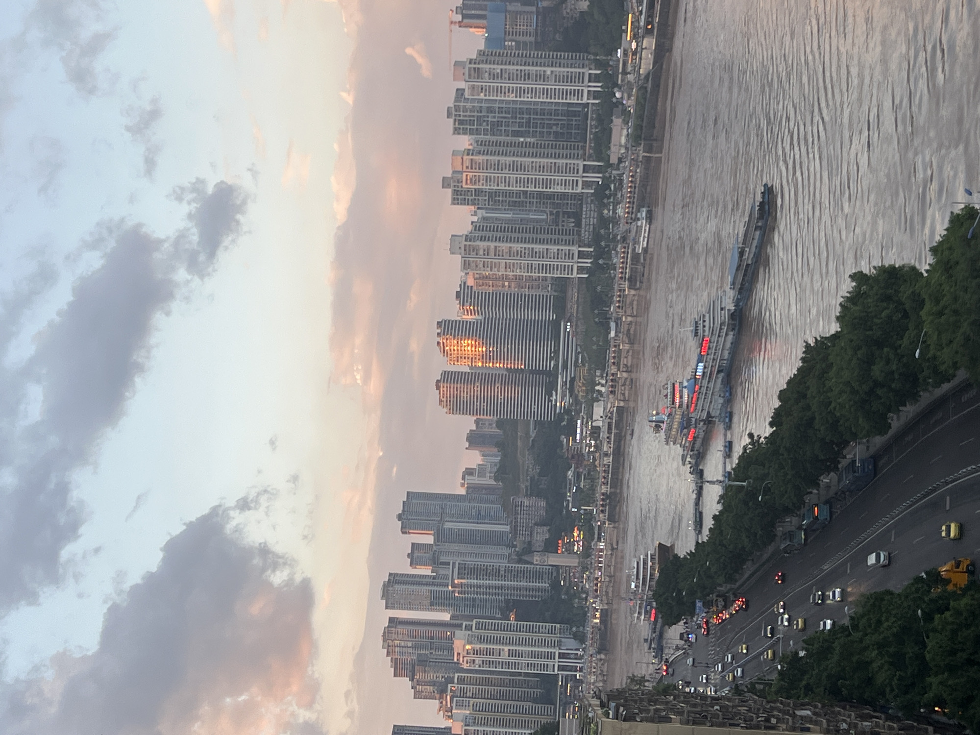 This is an aerial shot capturing a panoramic view of the cityscape and river in the Yuzhong District of Chongqing, China, during what appears to be late afternoon or early evening. The sky is a mix of soft blues, grays, and pastel pinks, with scattered clouds. Dominating the upper portion of the frame are numerous tall, modern residential buildings, densely packed and stretching across the horizon. The sunlight, catching the western facades of some of these buildings, creates a warm, golden glow.
Below the buildings, a wide, muddy river flows through the scene. Several boats are visible on the water. Two large passenger vessels, lit with red and white lights, are docked or moving slowly near the riverbank. Smaller boats and barges are also present.
Along the riverbank, a promenade or walkway is lined with trees, separating the water from a multi-lane road. Cars and buses are actively navigating this road, with headlights beginning to illuminate the traffic. The road curves and runs parallel to the river, bordered by lush green trees on one side and a concrete embankment or building structure on the other. The overall impression is a bustling urban environment integrated with its natural waterway.