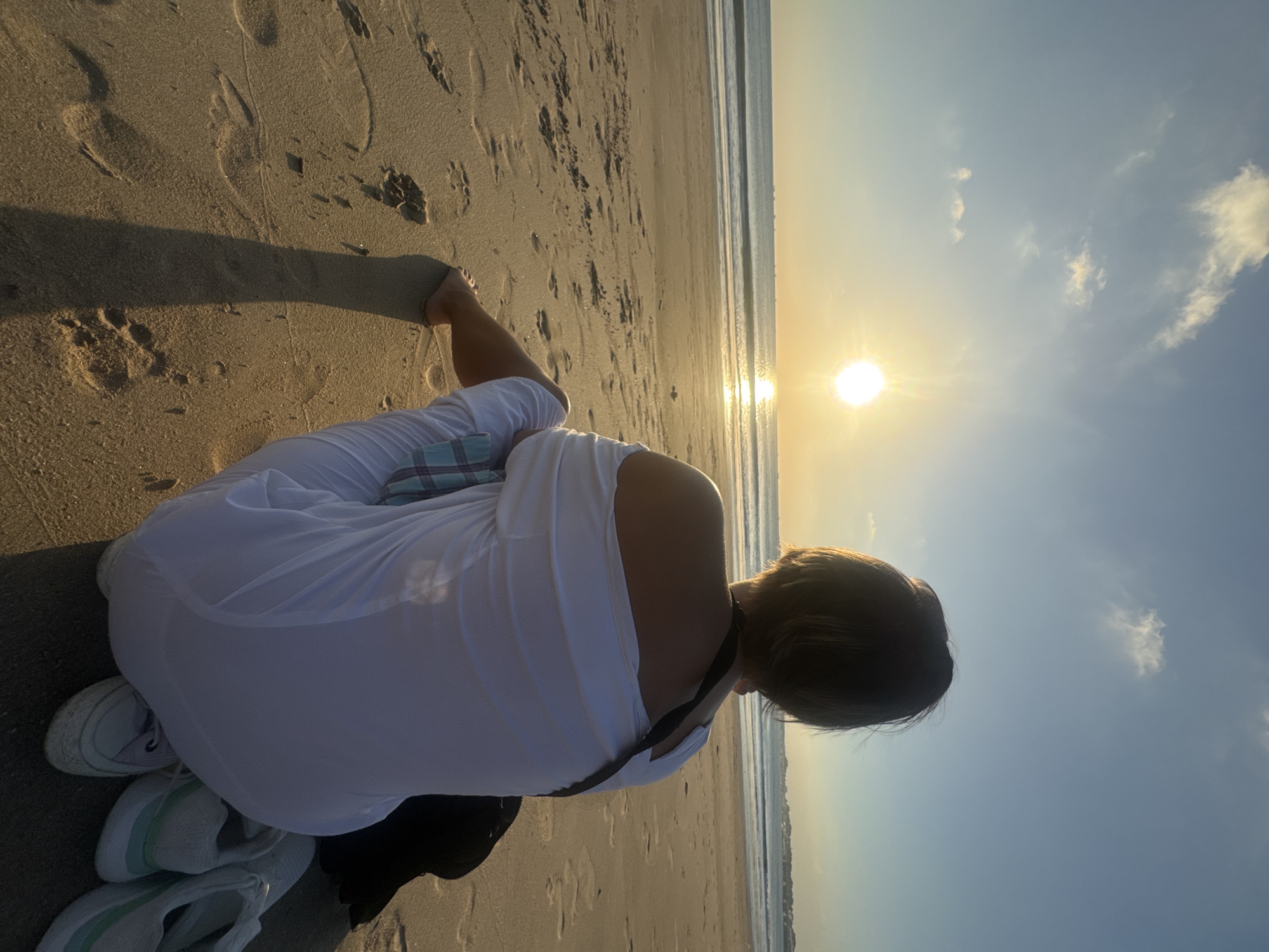 A person with short, dark hair sits on a sandy beach in Kuta, Indonesia, facing the ocean and the setting sun. The person is wearing a white off-the-shoulder dress and has a checkered bag strap across their shoulder. A pair of white sneakers with mint green accents are on the sand near the person. The sun is low in the sky, casting a warm glow and reflecting on the water. The beach is sandy with footprints visible. The ocean is calm with gentle waves and a distant horizon line. The sky is a clear blue with a few wispy clouds.