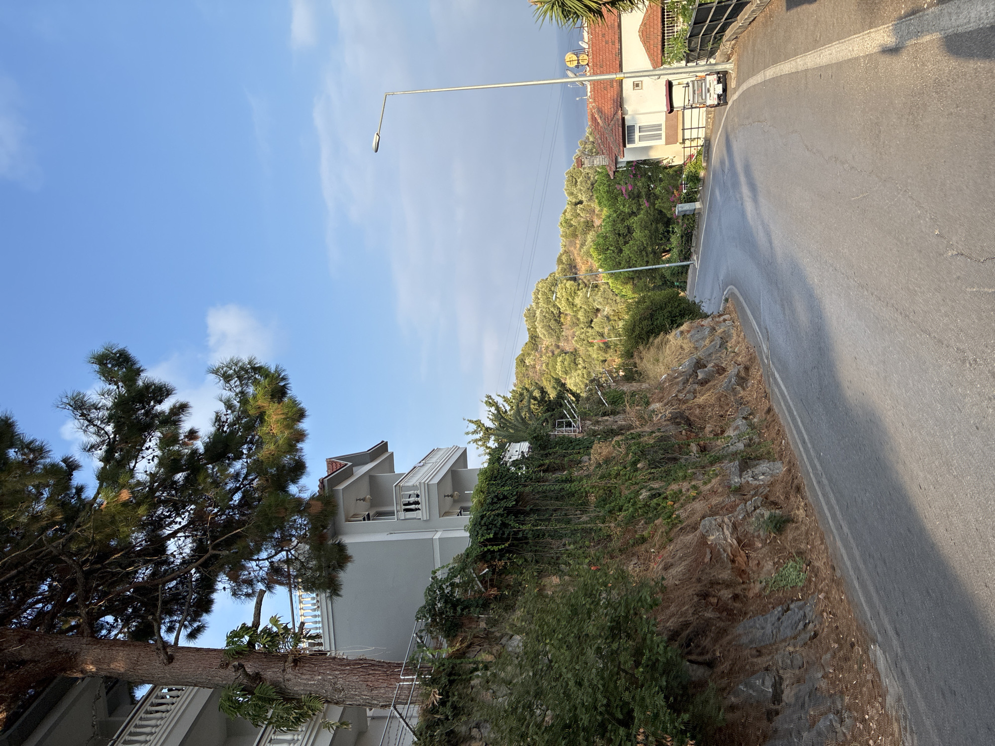 This is an image of a winding road in Alanya, Turkey, with a view of houses and lush greenery. The road is paved and appears to be a residential street. On the left side of the road, there is a steep, rocky embankment covered in vegetation, with a multi-story building perched on top. The building has white balconies and is partially obscured by a large pine tree. On the right side of the road, there are houses with red-tiled roofs. One house has a small, white vehicle parked in front of it. In the background, there are rolling hills covered in trees, and a glimpse of the sea can be seen. The sky is blue with a few scattered clouds. The lighting suggests it is either early morning or late afternoon, with long shadows cast across the road.