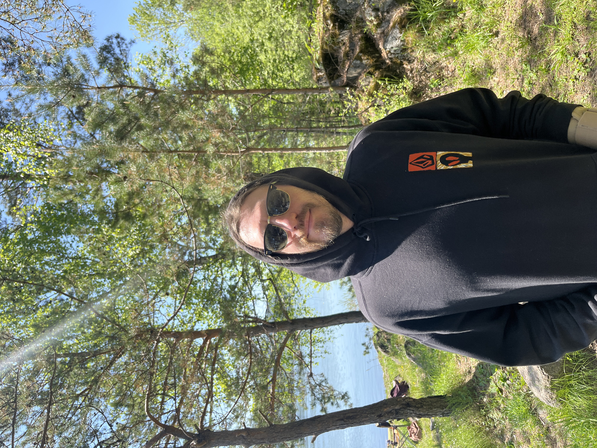 A man wearing a black hoodie with the hood up and sunglasses gazes directly at the camera in a vibrant outdoor setting. He has a beard and his expression is neutral to slightly smiling. On the front of his hoodie, a stylized, multi-colored logo featuring red, orange, and gold shapes is visible. He is positioned near the grassy bank of a calm body of water, likely a lake or wide river, which stretches into the background reflecting the blue sky.
The surroundings are lush with greenery, indicating a natural, forested environment. Tall trees with green foliage and pine needles dominate the background, with a clear blue sky visible through the branches, suggesting bright, sunny weather during the daytime. On the grassy bank in the foreground, what appears to be fishing equipment, including fishing rods on stands and a bag, is set up, implying that fishing is the activity taking place. The scene depicts a peaceful, sunny day in a natural outdoor location, specifically near Sliznevo, Russia.